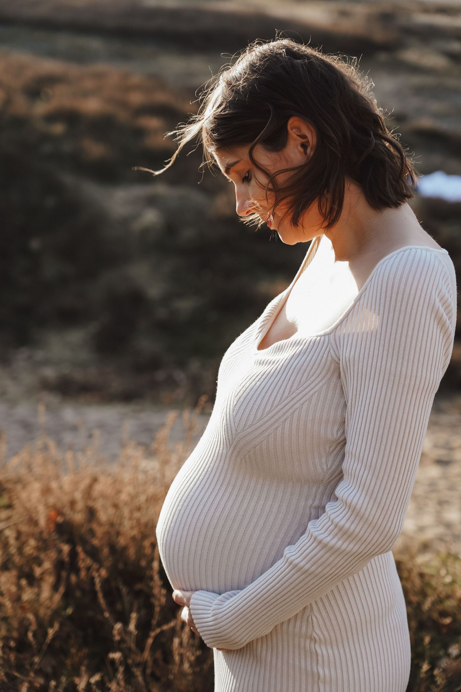Side view of expecting mother in white ribbed dress during golden hour outdoor photoshoot.