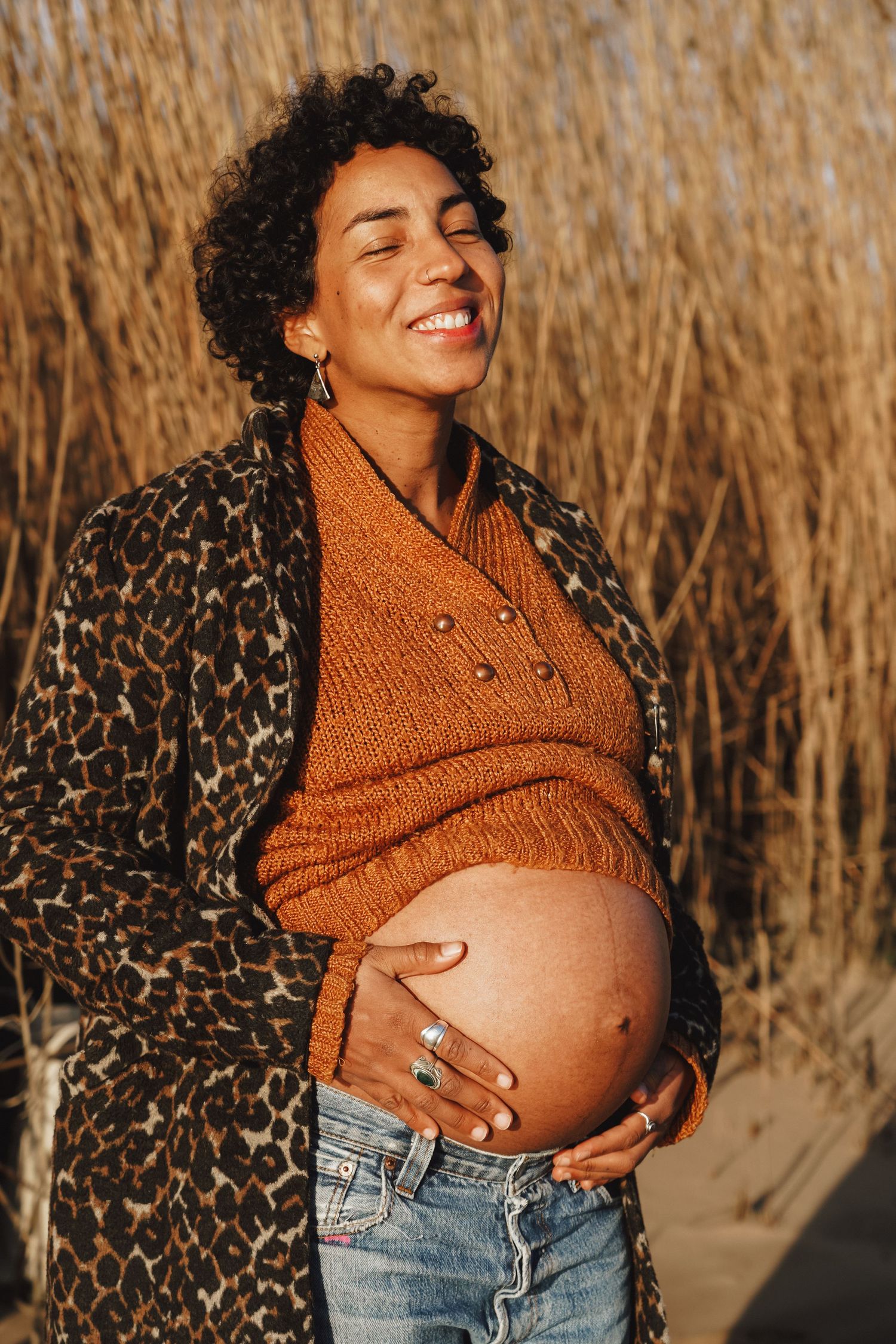 A maternity portrait in a leopard print coat and orange sweater against tall grass at sunset.