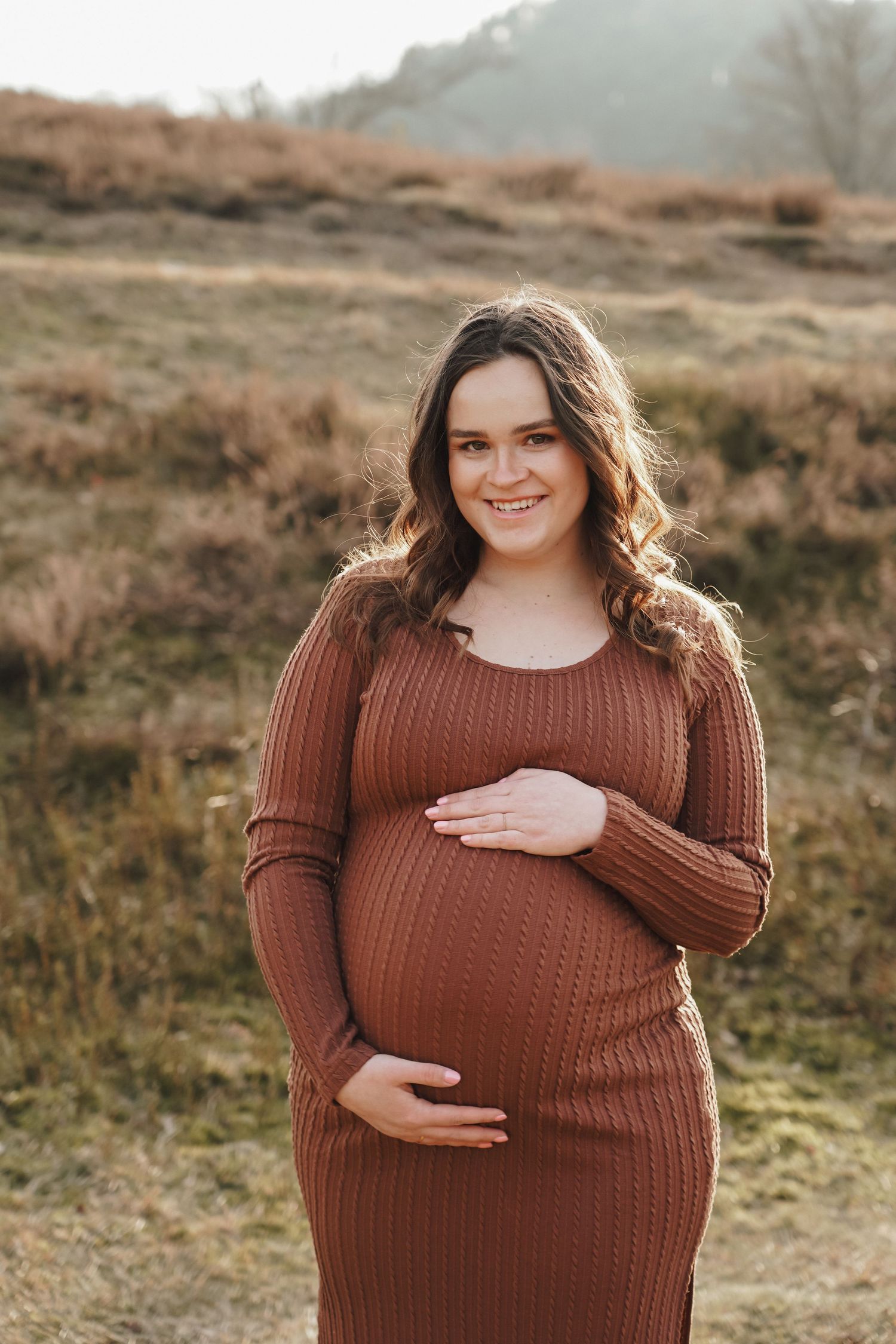 A maternity portrait in a brown sweater dress standing in a sun-lit field during golden hour.