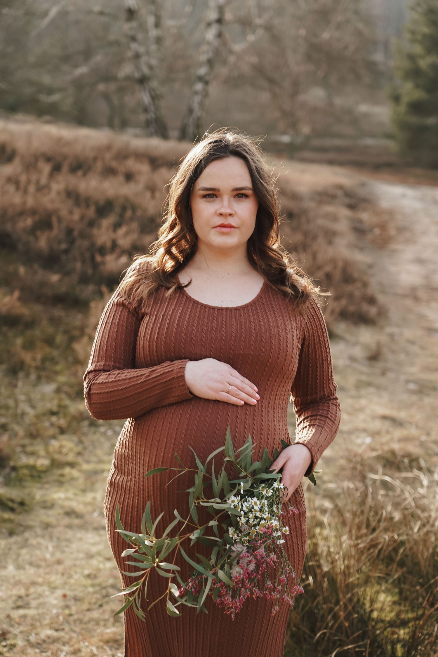 A maternity photo in autumn featuring a brown ribbed dress and wildflower bouquet against a natural backdrop.