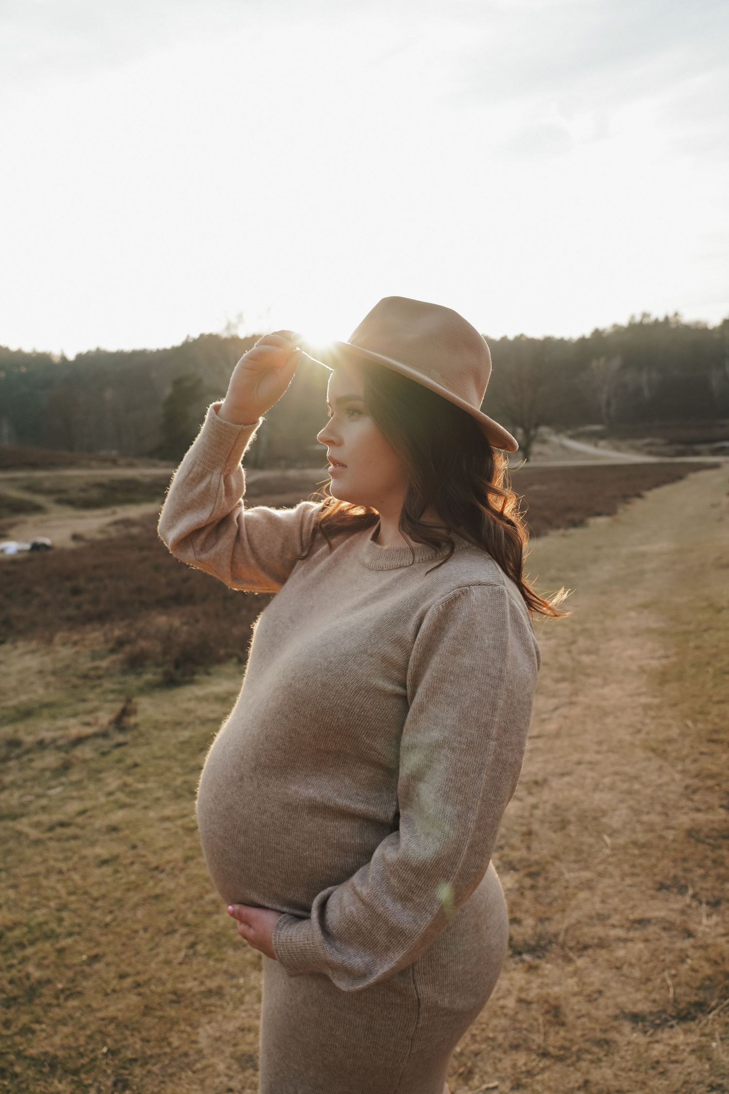 A maternity portrait at golden hour featuring a grey dress and brown fedora in a natural outdoor setting.