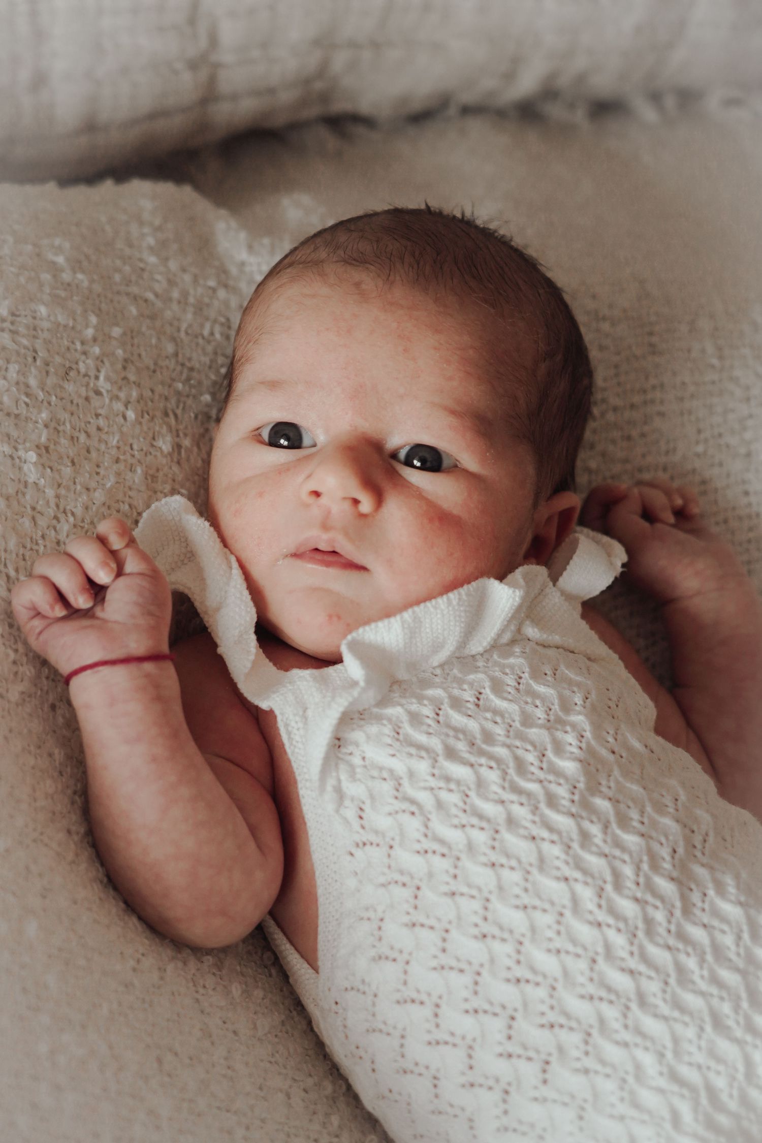Newborn baby wearing white knit clothing lying on soft beige fabric.