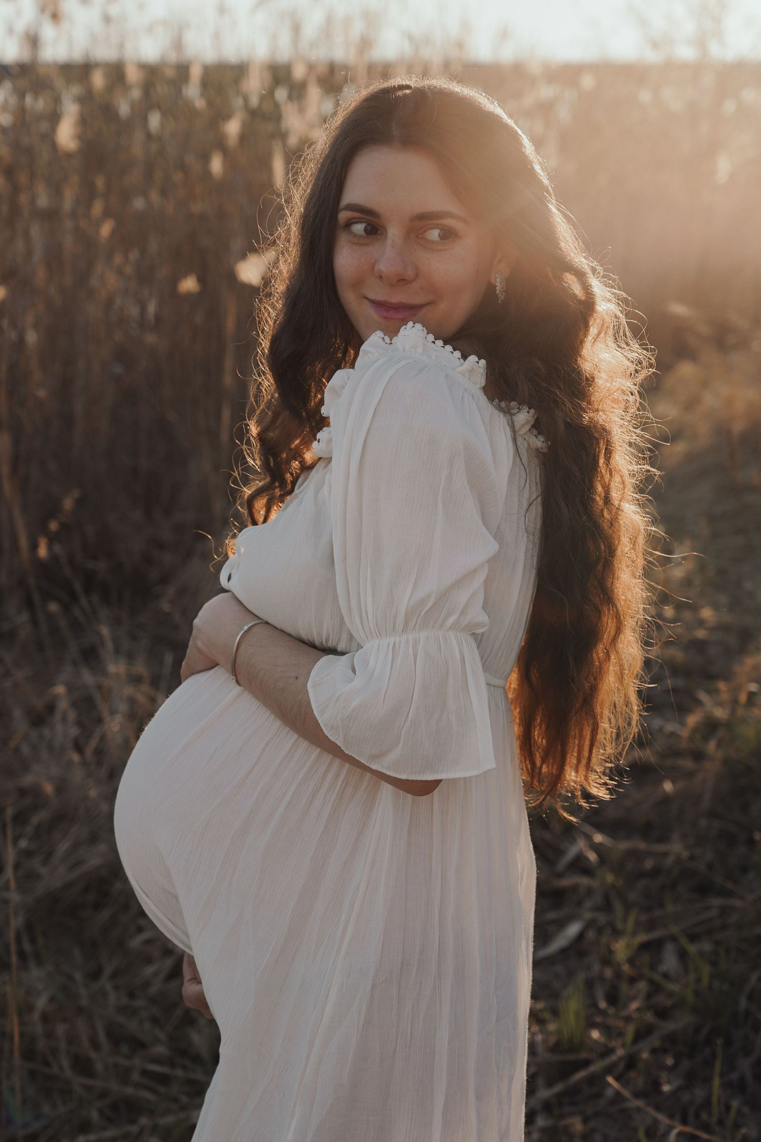Beautiful maternity photo of a woman in a flowing white dress at golden hour with sunlight creating a dreamy backlit glow.