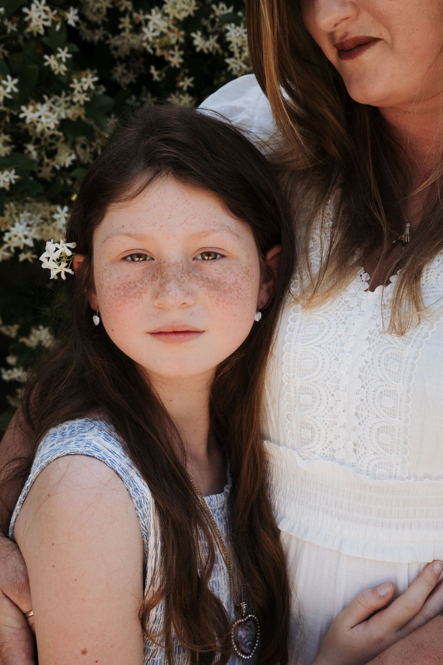 Close up portrait of a young girl with beautiful eyes staring into the camera as her mom holds her near. Girl has a flower in her ear.