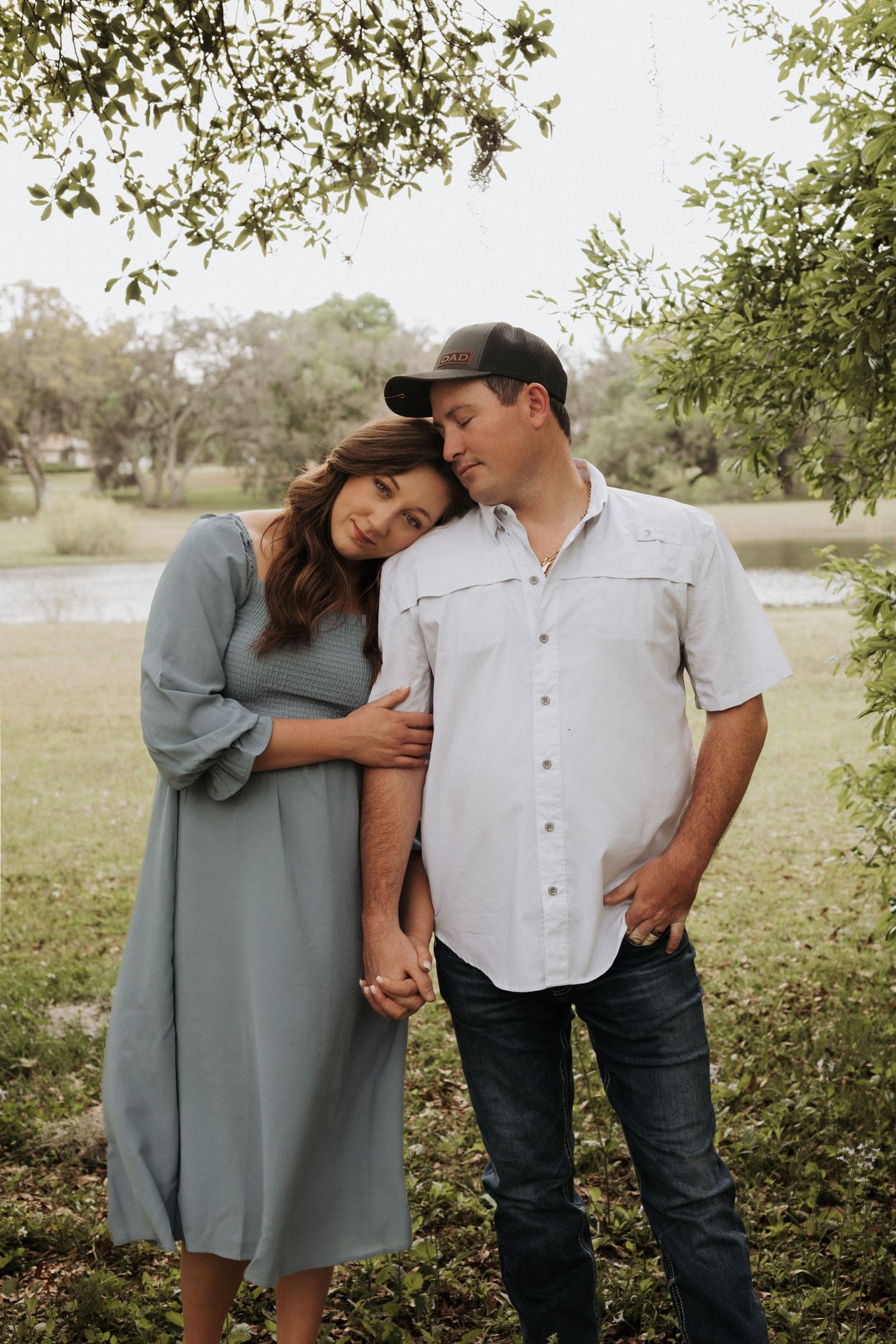 Portrait of couple in a nature setting. Wife is resting her head in husbands's shoulders as he is holding her hand facing her.