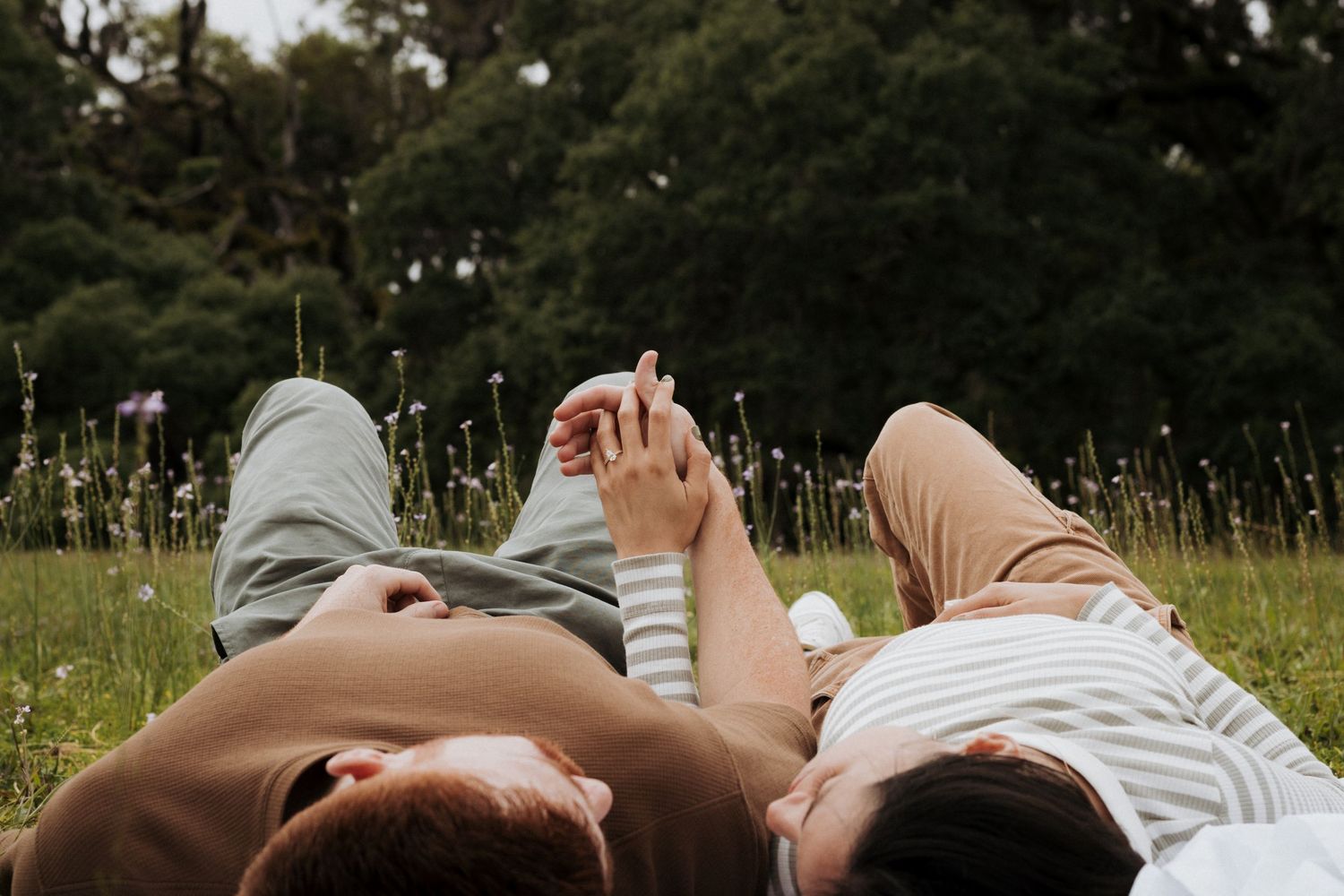 Lifestyle portrait of couple laying down in a field of small flowers, looking at each other while holding hands.