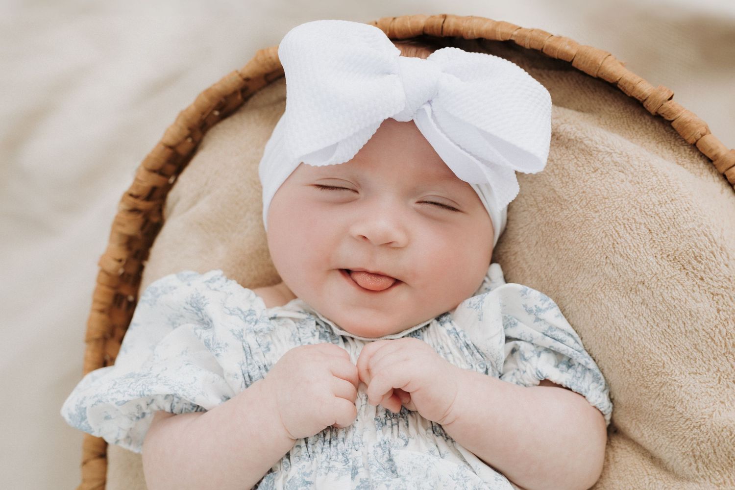 Portrait of a baby in a basket wearing a white bow and she is smiling and sticking her tongue out.