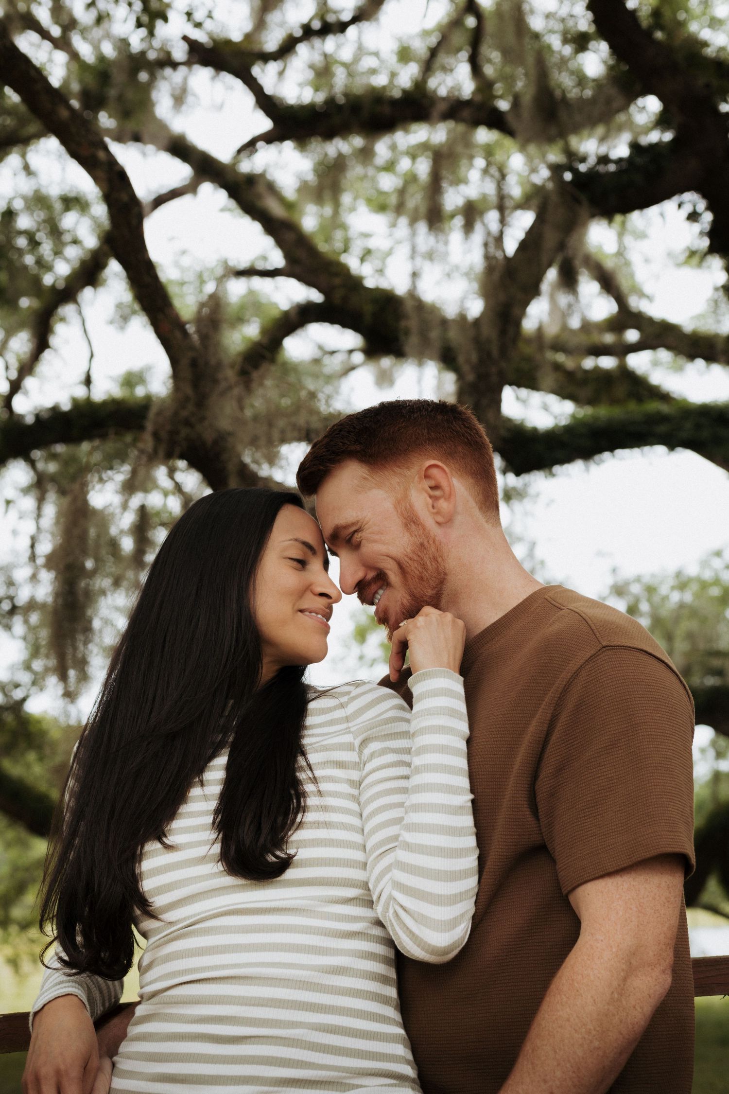 Couple portrait with oak tree branches in the background. Female is leaning on man holding his chin, male is leaning toward her and smiling.
