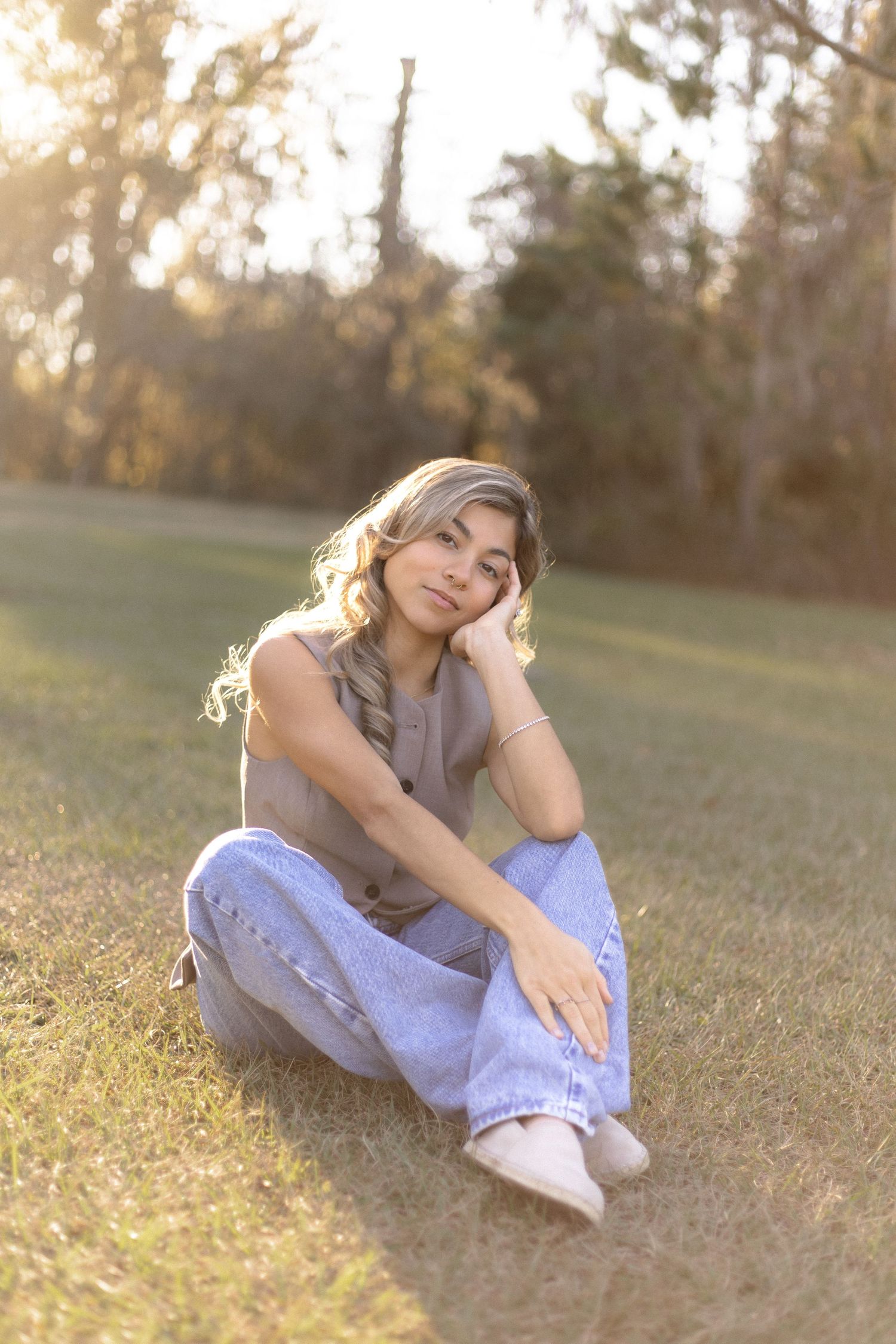 Young female portrait during golden hour in a grassy field as she is sitting with her legs crossed and one hand holding her face.