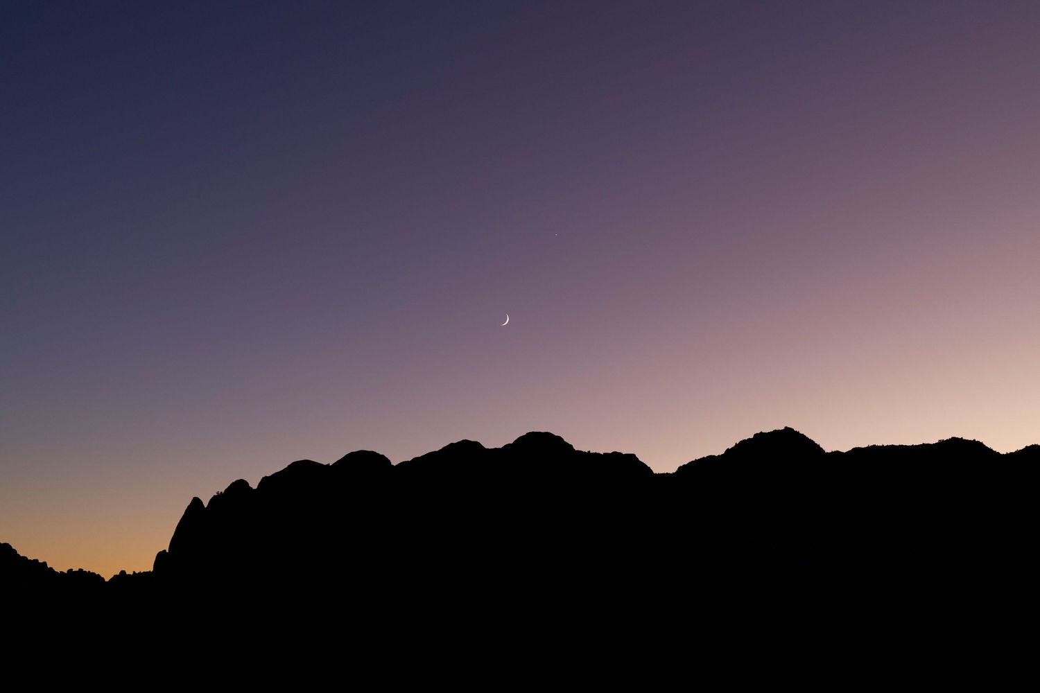 Purple skies as sun has just set behind a mountain. Showcasing the mountain as a silhouette to focus on colors of the sky & crescent moon.