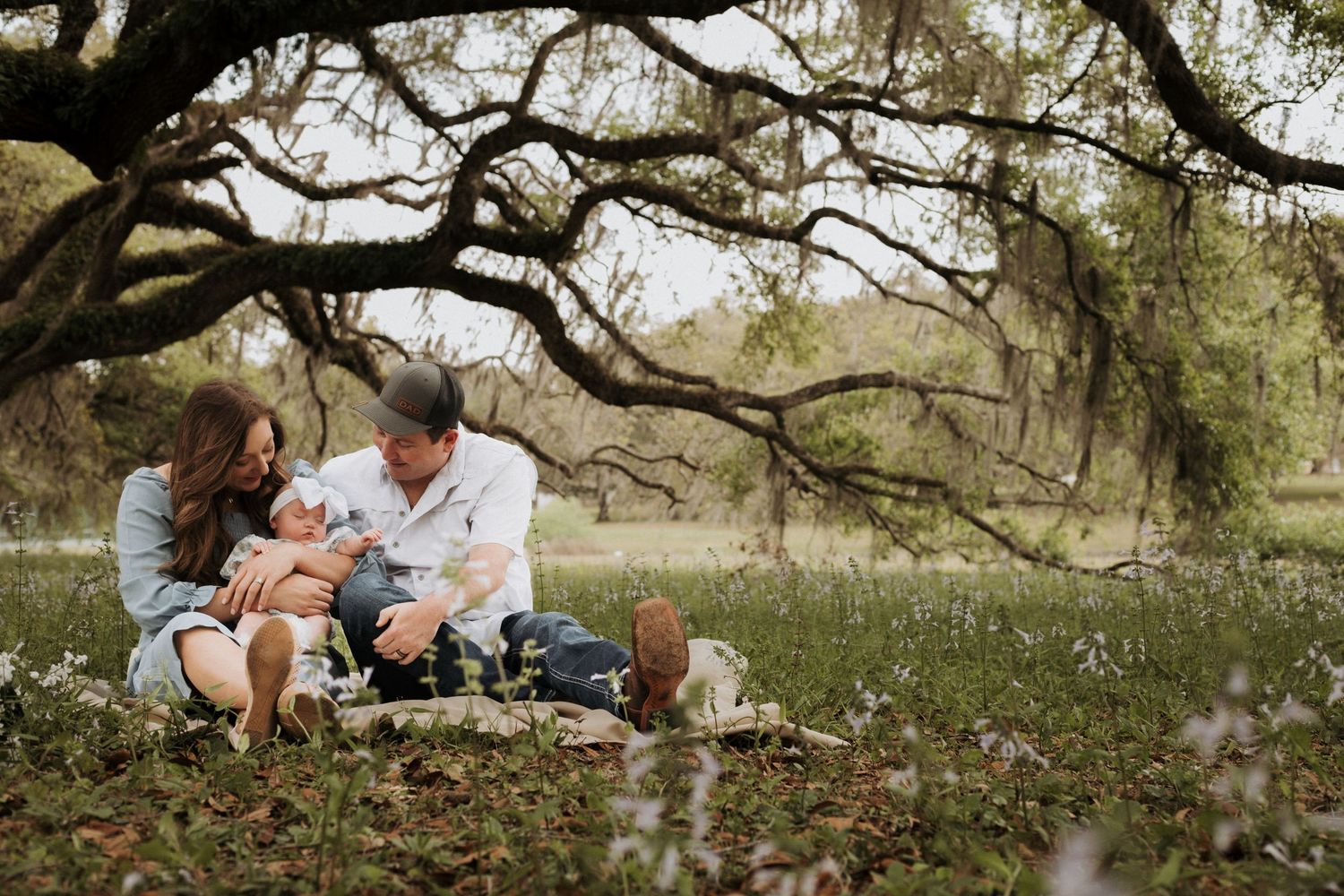 Portrait of family in a nature setting sitting on a blanket holding their babygirl while they're looking at her and smiling.
