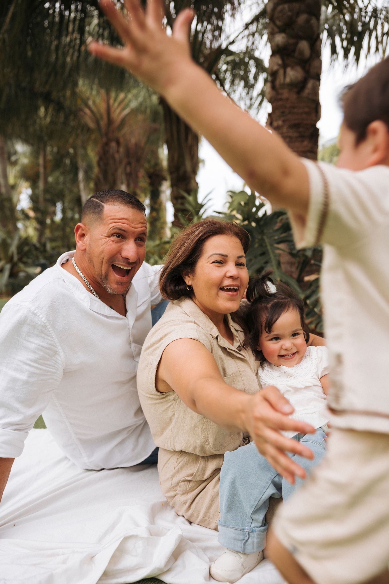 Action portrait of a toddler boy running towards his grandparents who are sitting in a white blanket holding another girl, all smiling.