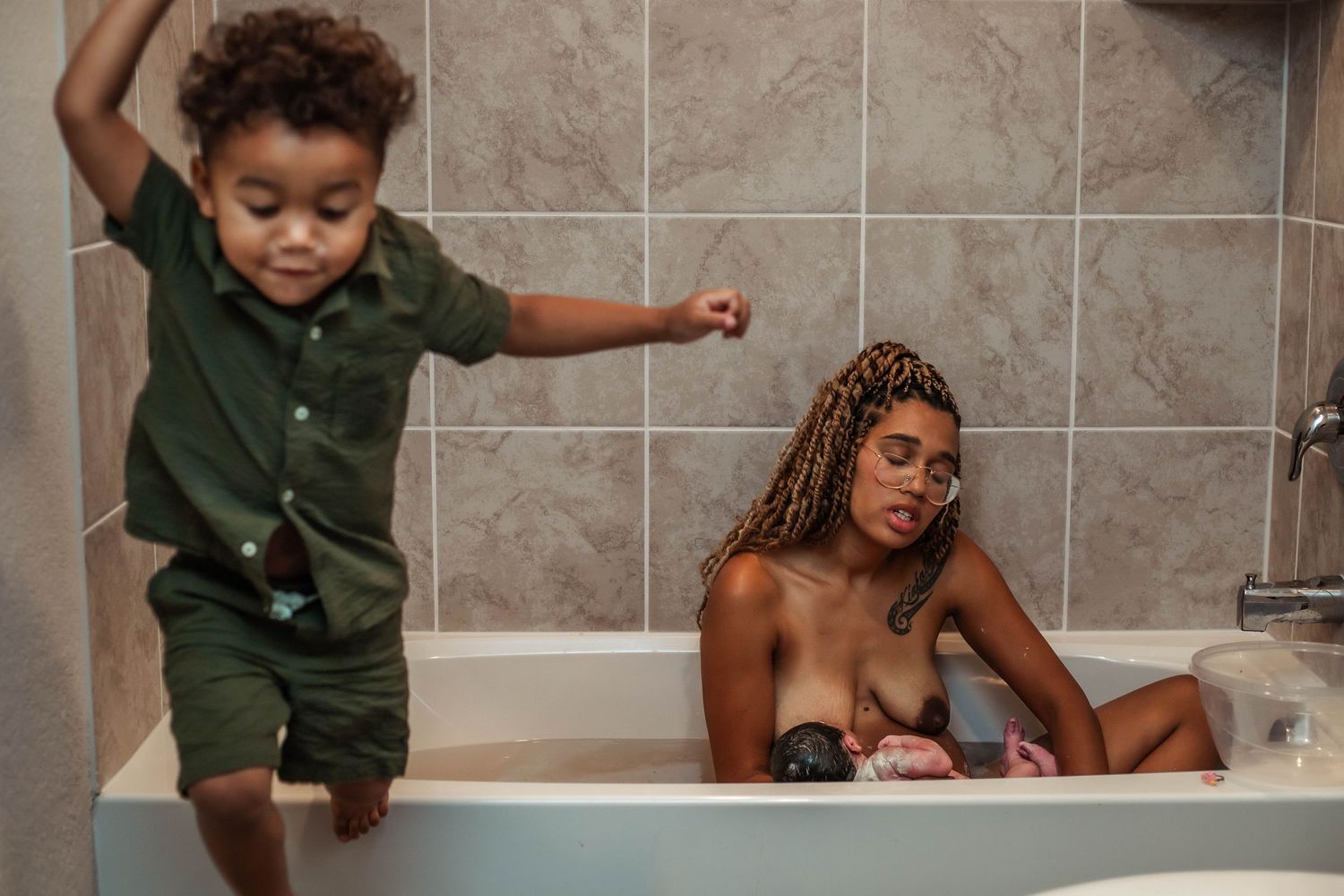 Mother holds her newborn baby after birth nursing in home bathtub while older child jumps from the edge of the tub.