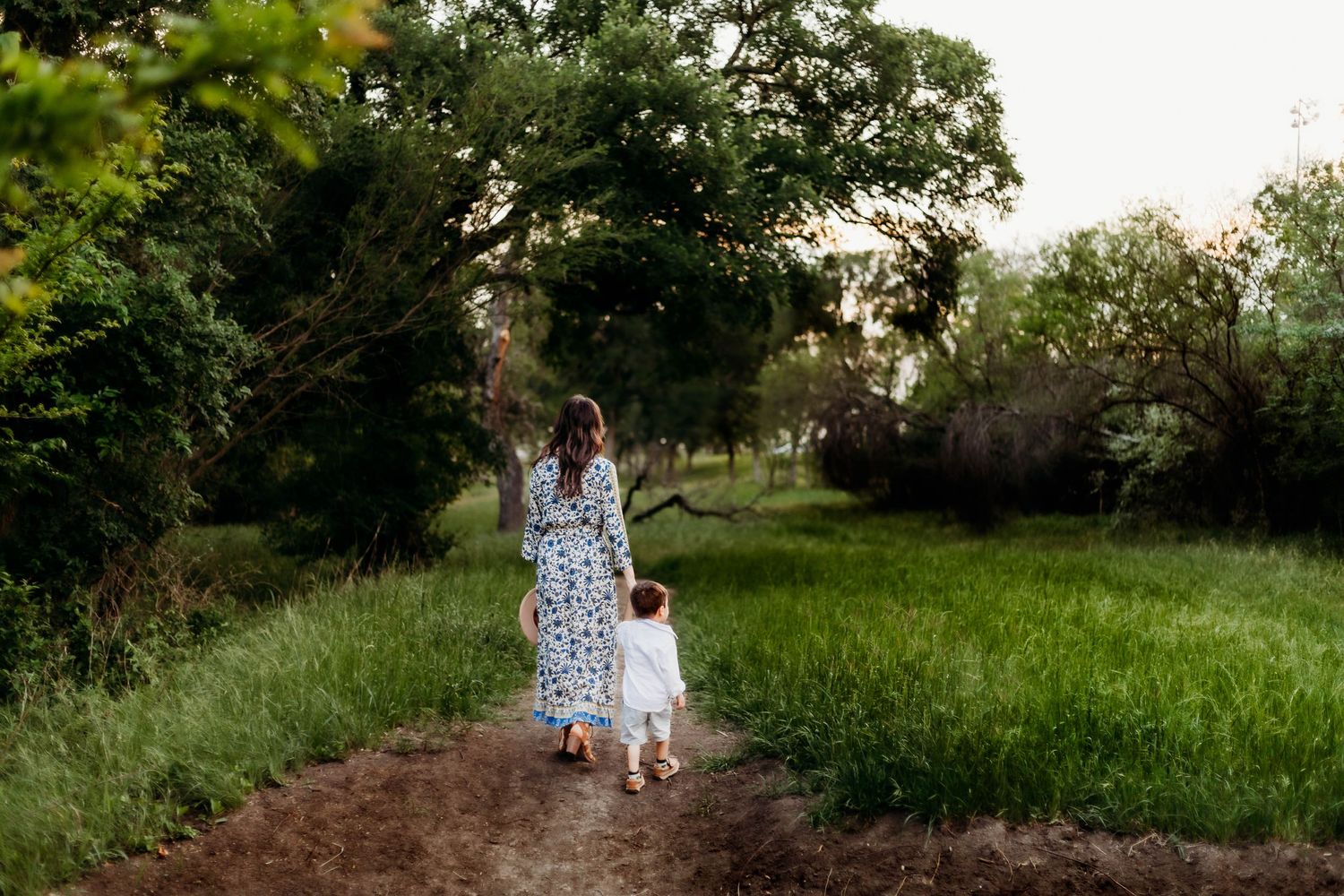 Mother and son walk down a dirt road surrounded by trees and a green field