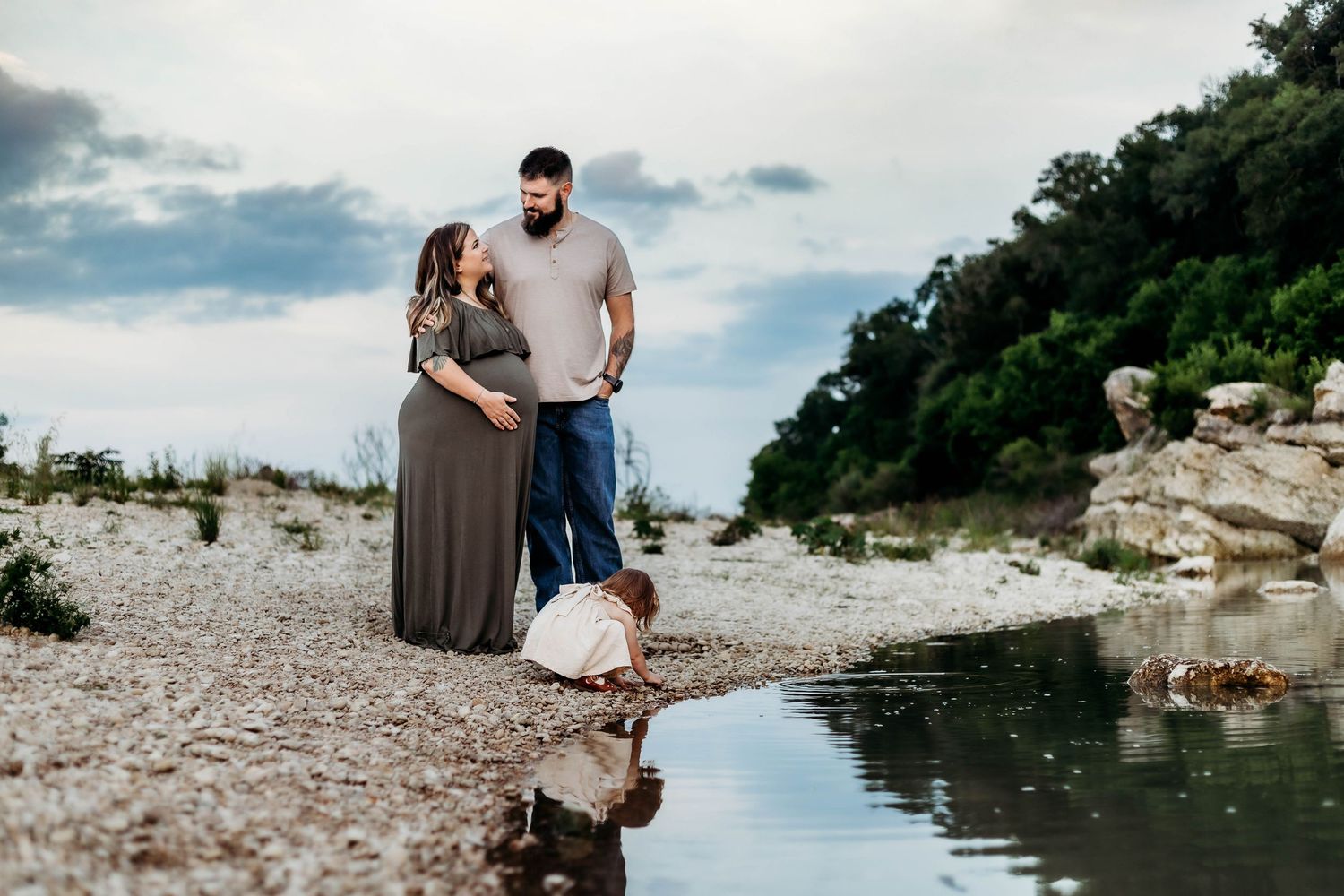 Pregnant mother smiles at her husband while their older daughter plays at their feet next to water