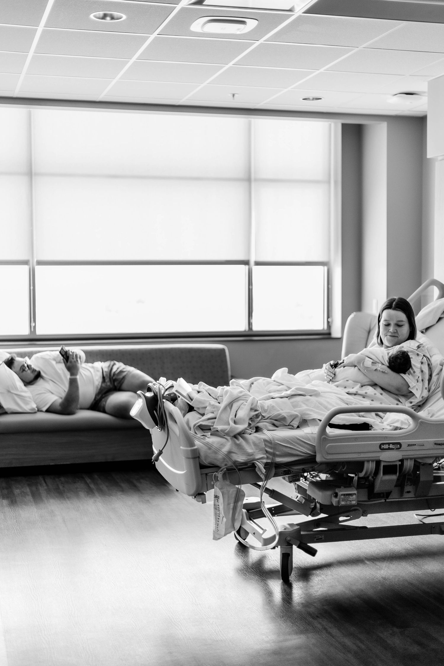 Full view of hospital room, mother holds and looks at baby, while father lays on couch on his phone