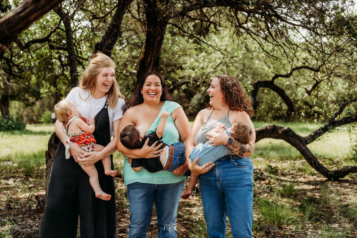 Three moms stand laughing together while nursing their little ones during a breastfeeding awareness shoot