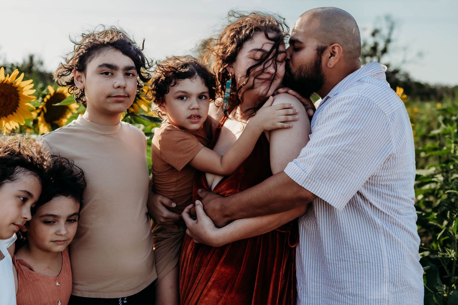 Family poses in front of sunflowers, husband kisses wife's head, while she holds their little boy, standing next to her are her older kids