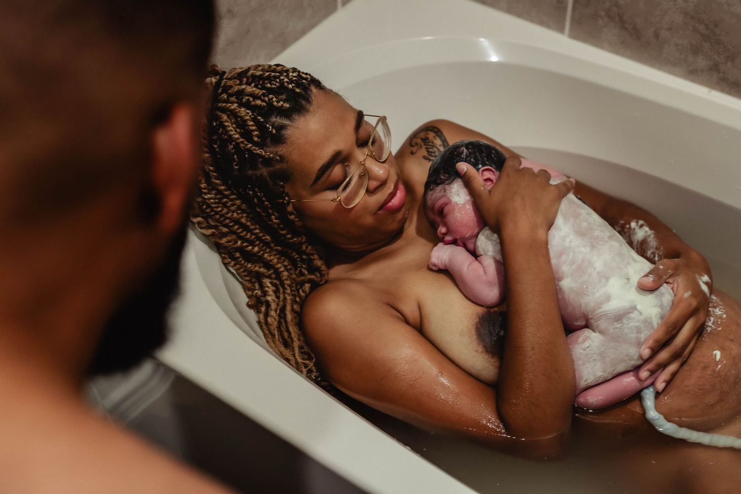 Mother holds newly born baby while husband looks at them in their home bathtub.