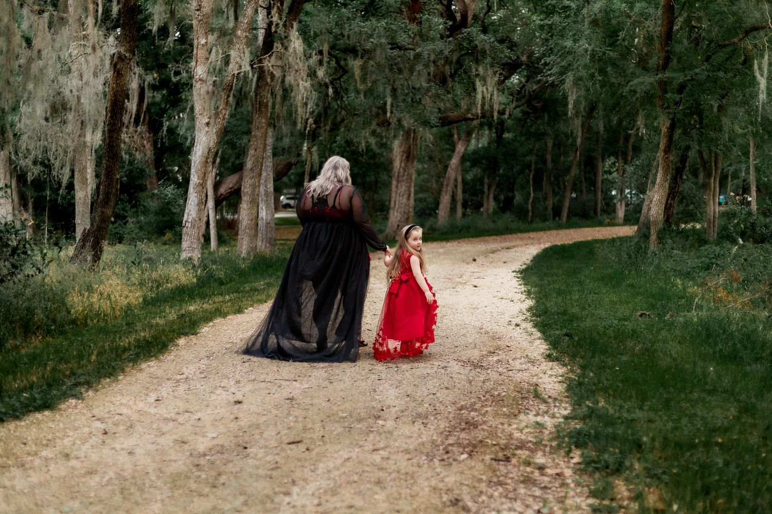 Mother and daughter walk down a dirt road surrounded by green trees, daughter looks back