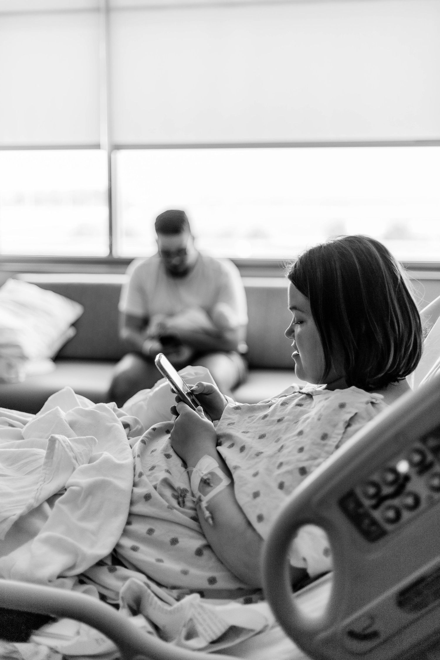 Mother checks her phone while father holds baby in the background in hospital room