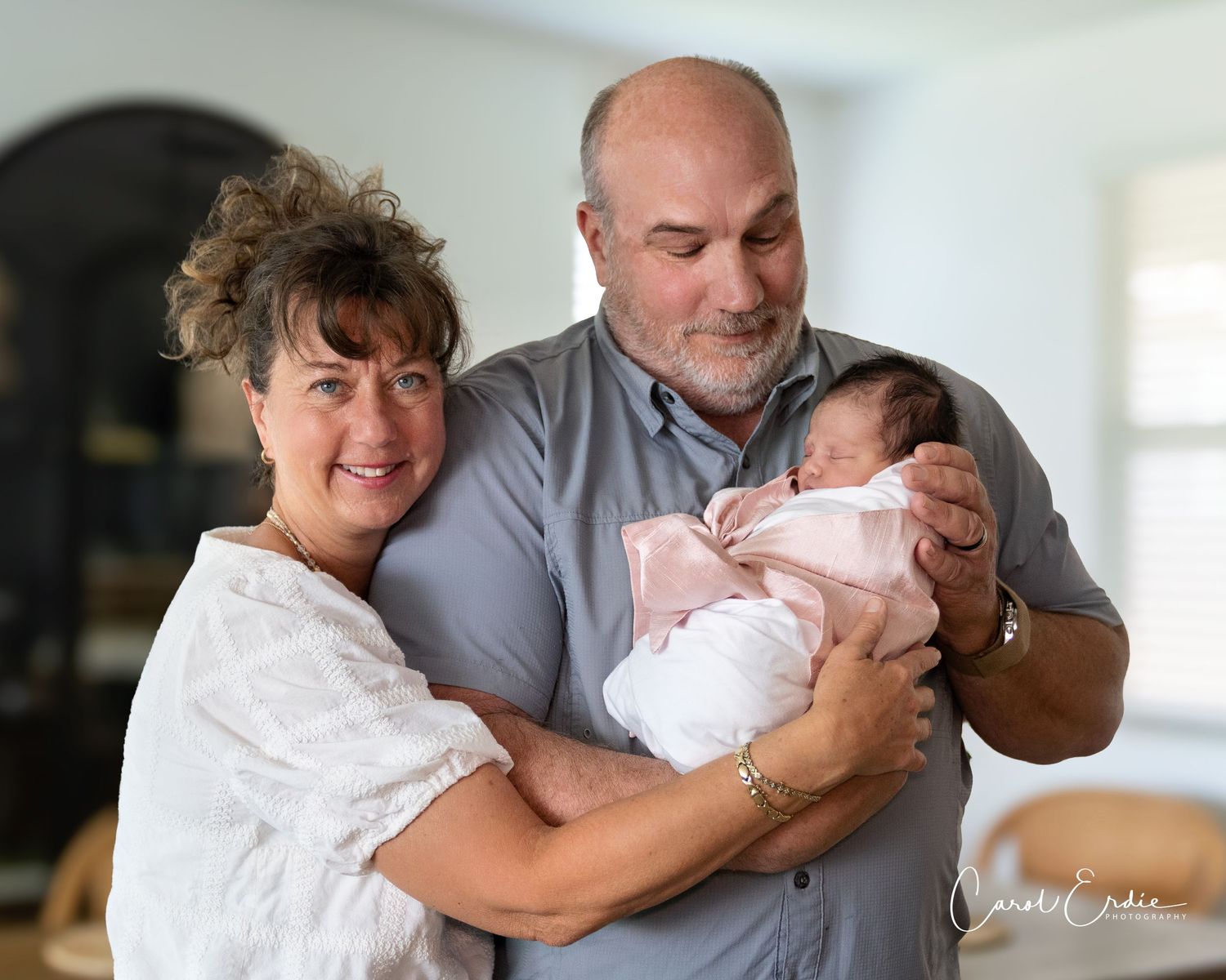 Grandparents smiling while holding their newborn grandchild wrapped in a pink blanket indoors in Phoenix, AZ.