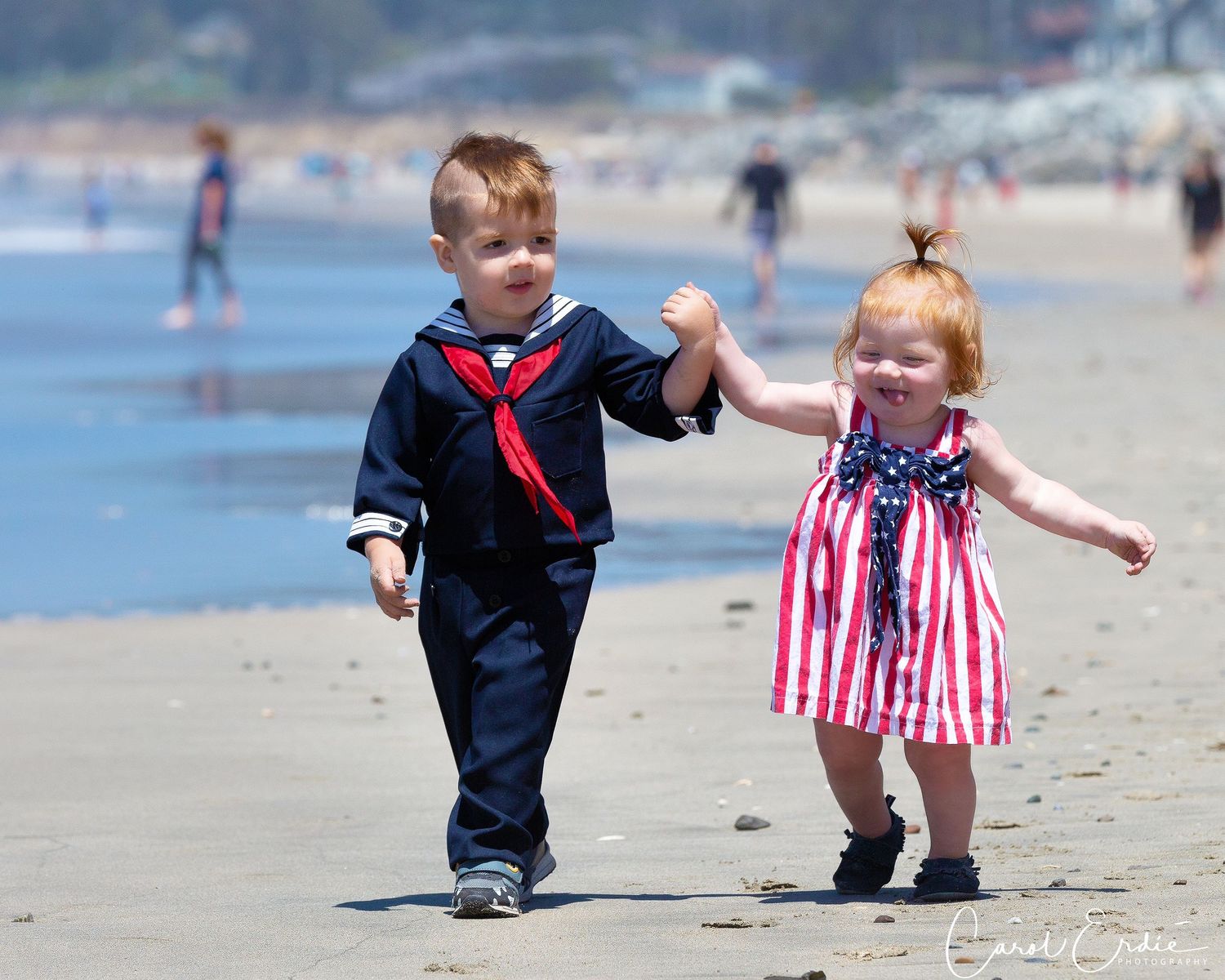 Toddlers in patriotic outfits playing on a sandy beach in Half Moon Bay, CA, with ocean waves in the background.