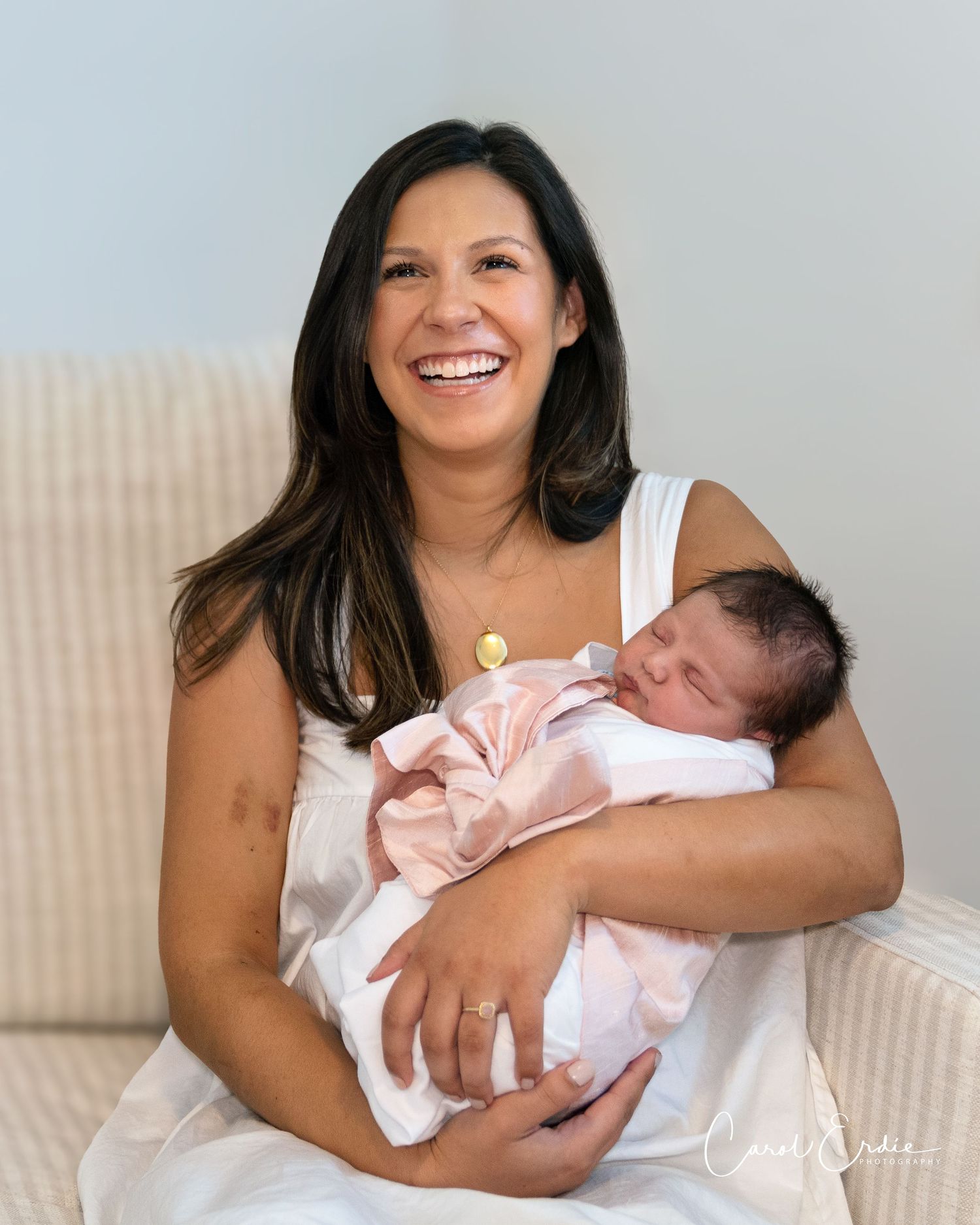 Smiling mother in a white dress holding her newborn baby wrapped in a pink blanket, sitting indoors in Phoenix, AZ.