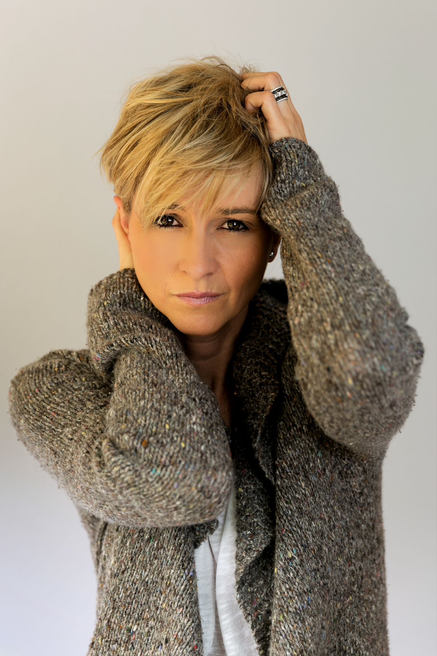 Portrait of a woman with short blonde hair wearing a gray knitted sweater, posing with hands on her head.