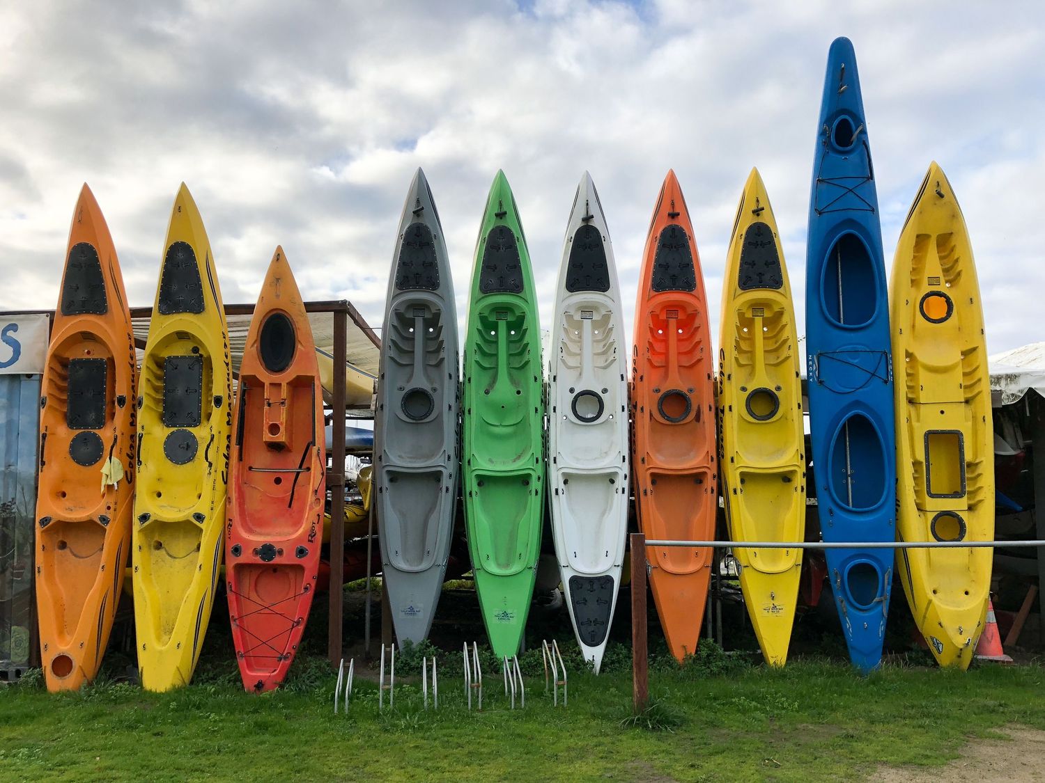 A row of colorful kayaks standing upright on grass with a cloudy sky background in Half Moon Bay, CA.