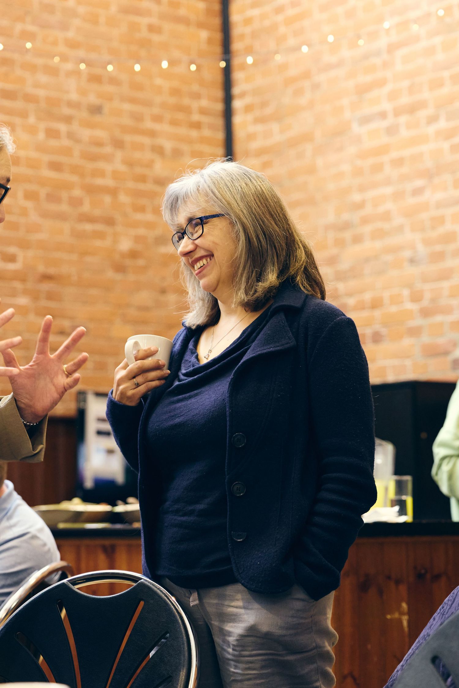 Smiling older woman with glasses holding a cup, wearing a navy blazer, chatting in a brick-walled venue.
