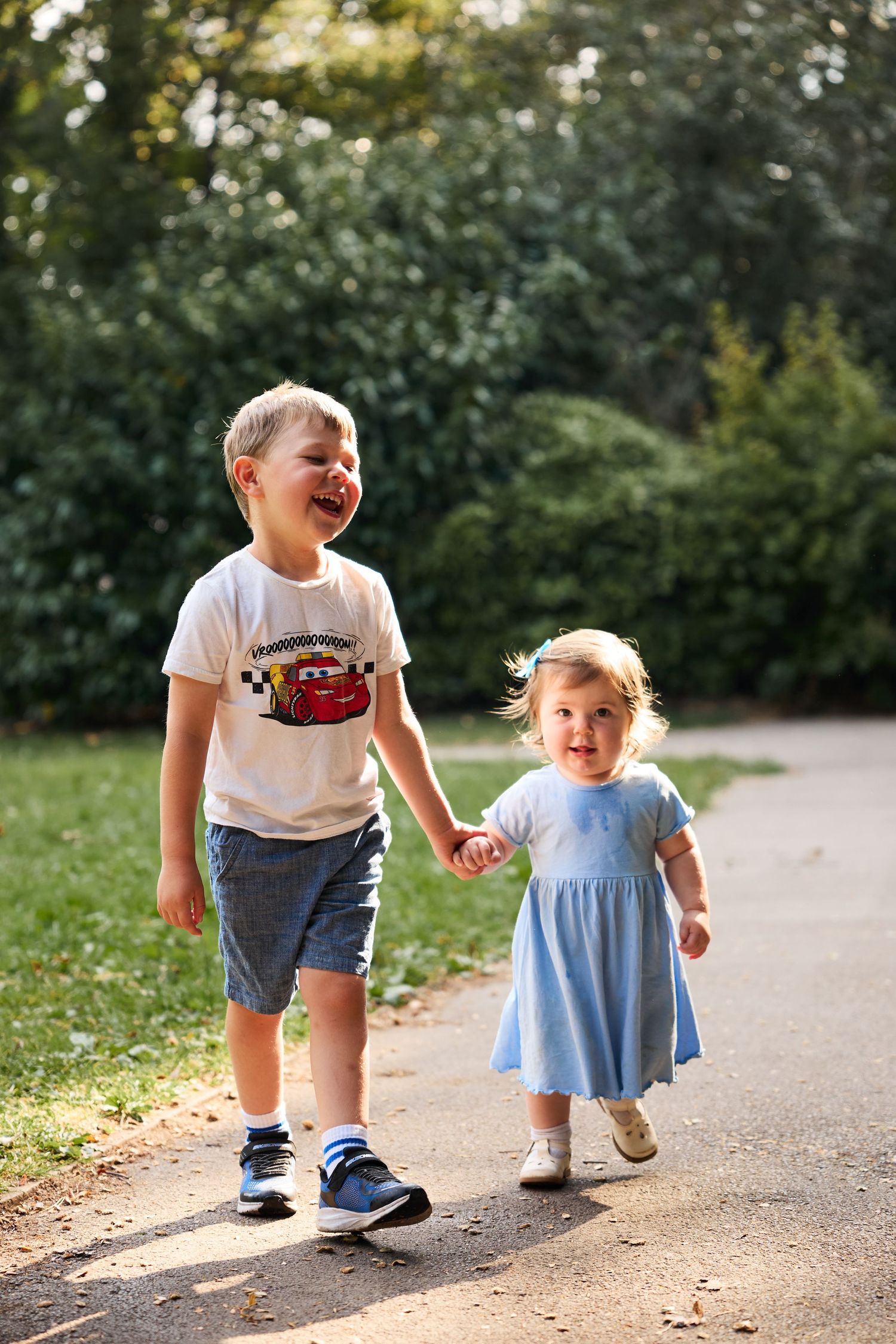 Children in casual wear laugh and play together on a paved path during a sunny day.