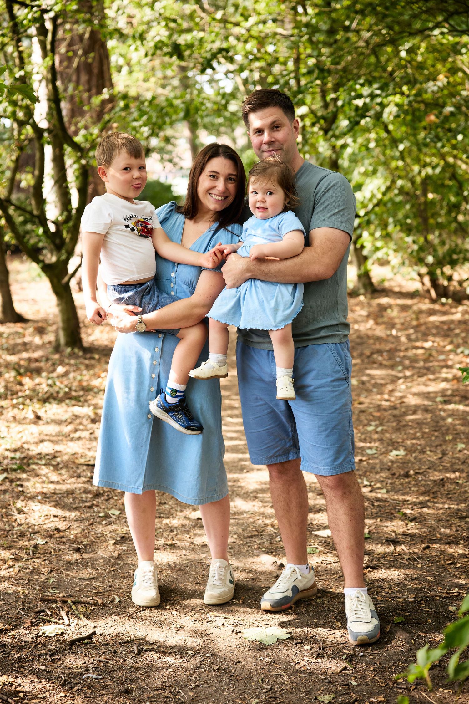 Family members wearing coordinating blue outfits stand together on fallen leaves in a wooded area.