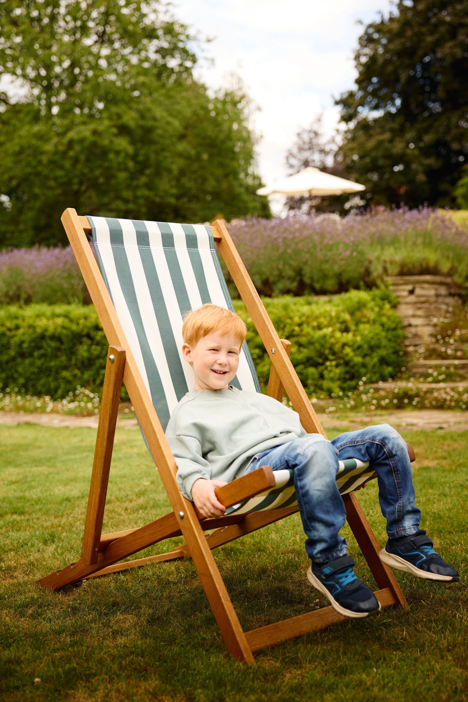 Child relaxing in a striped deck chair on a sunny lawn with garden in background.
