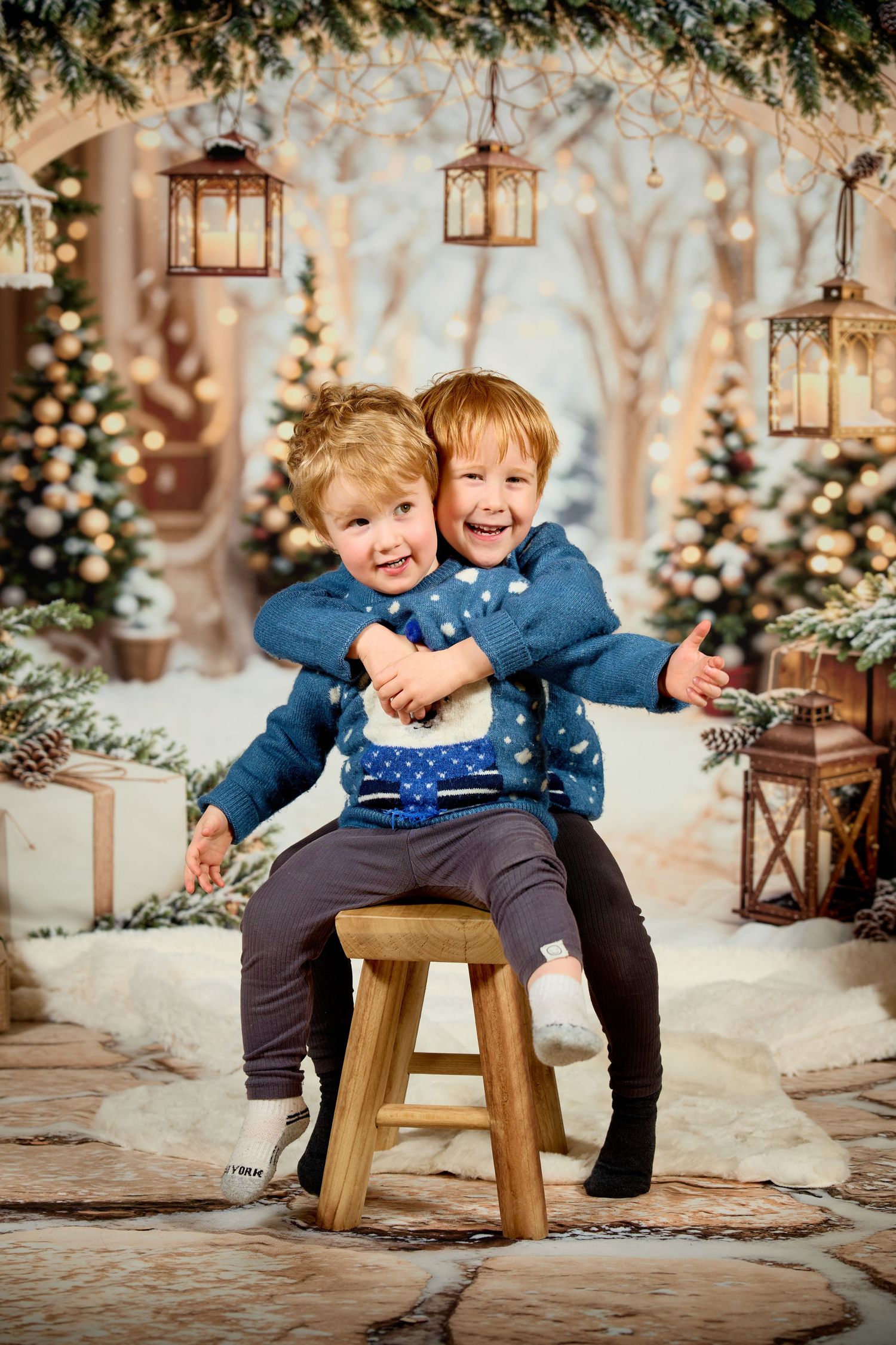 Two children in matching blue sweaters playfully pose together on a wooden stool in a winter wonderland setting.