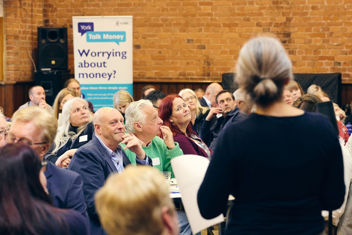Audience attending a financial wellness seminar with a Talk Money banner asking Worrying about money? in a community hall.
