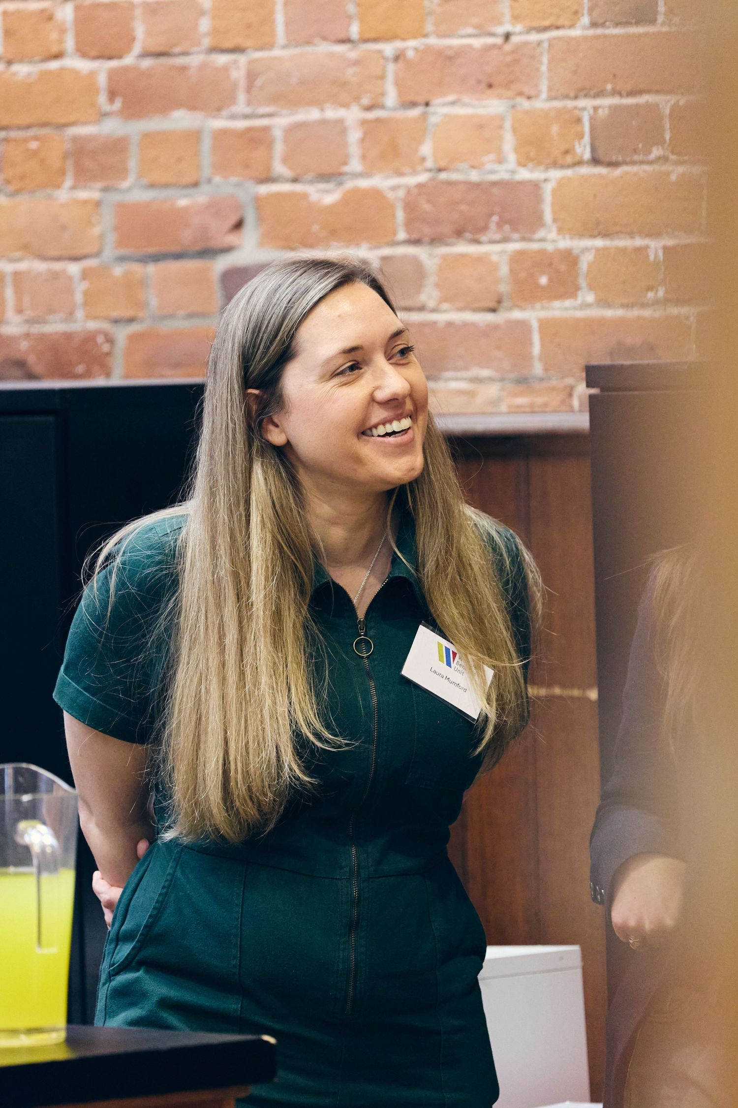 Smiling woman in green dress with name tag laughing at an indoor event near exposed brick wall.