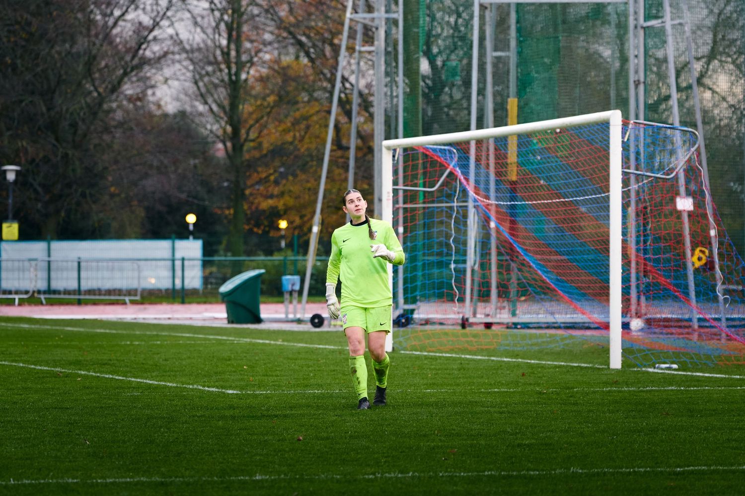 Soccer player in bright yellow goalkeeper uniform prepares to defend goal during outdoor match on grass field.