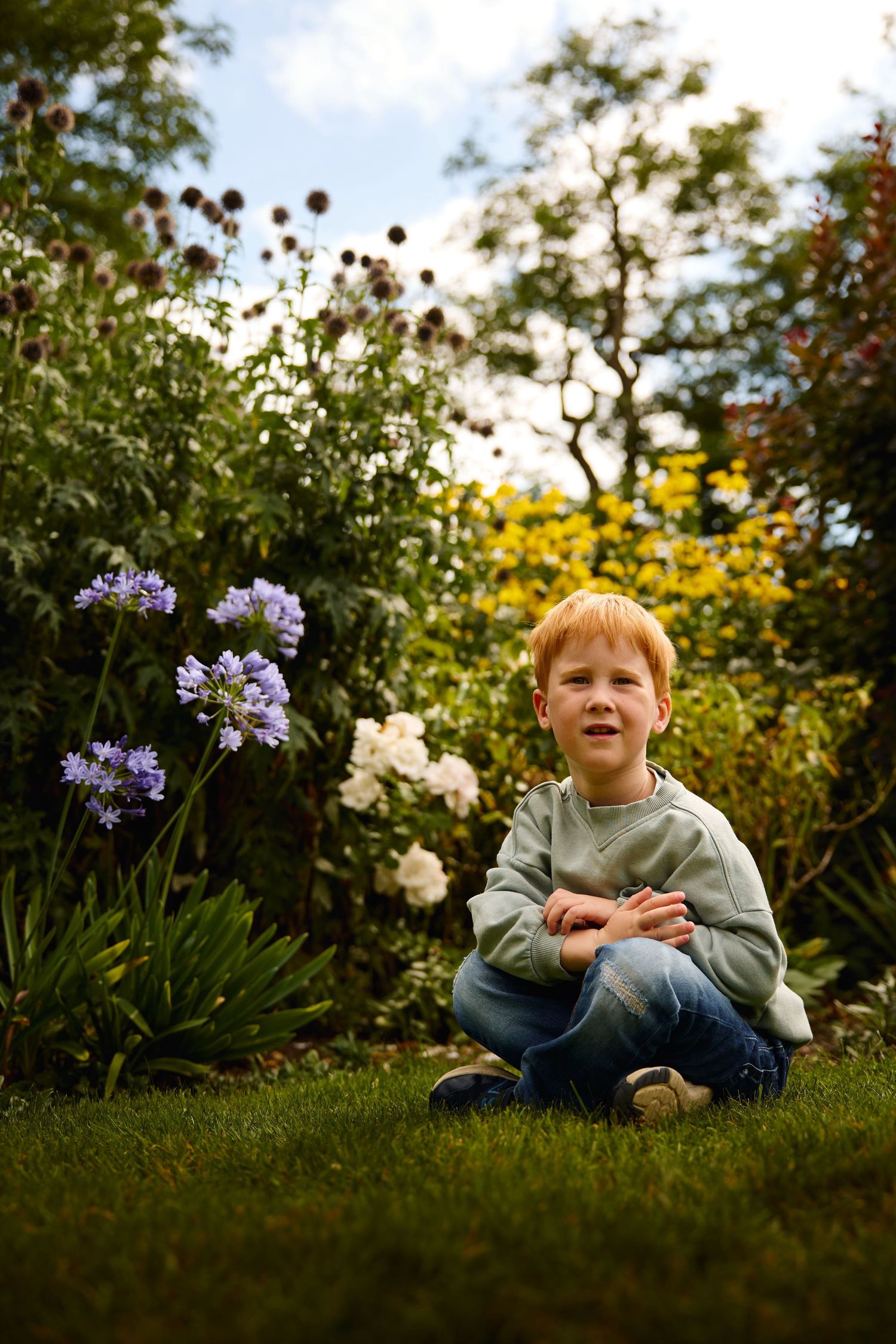 Child sitting on grass surrounded by blooming purple and yellow flowers in garden setting.
