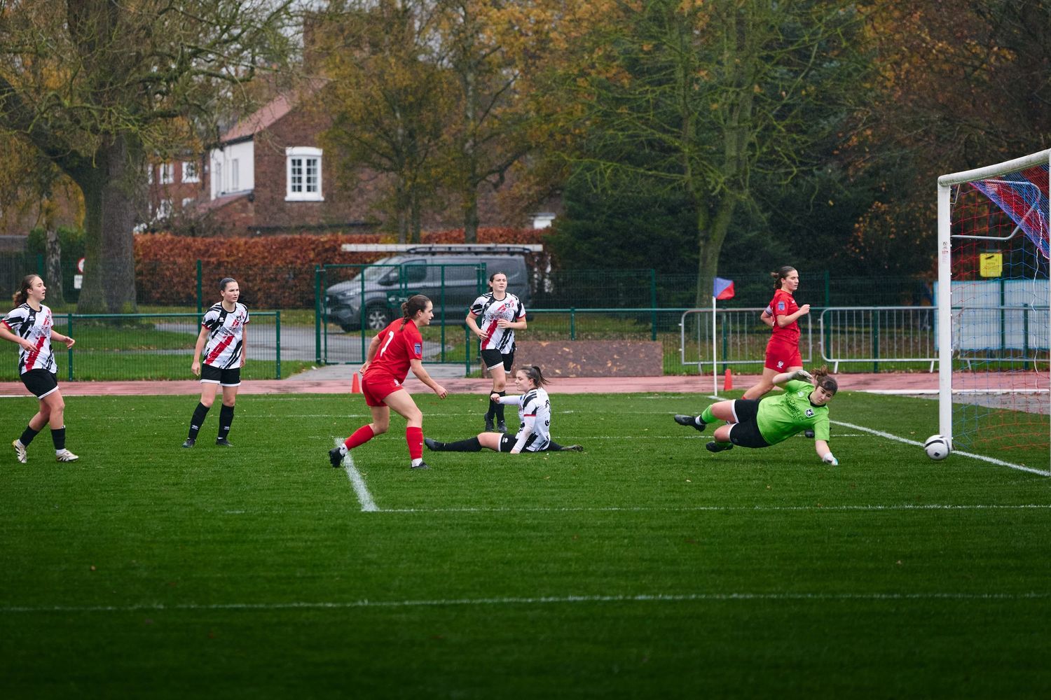 Soccer players compete in a match on a grass field surrounded by autumn trees during a cloudy day.