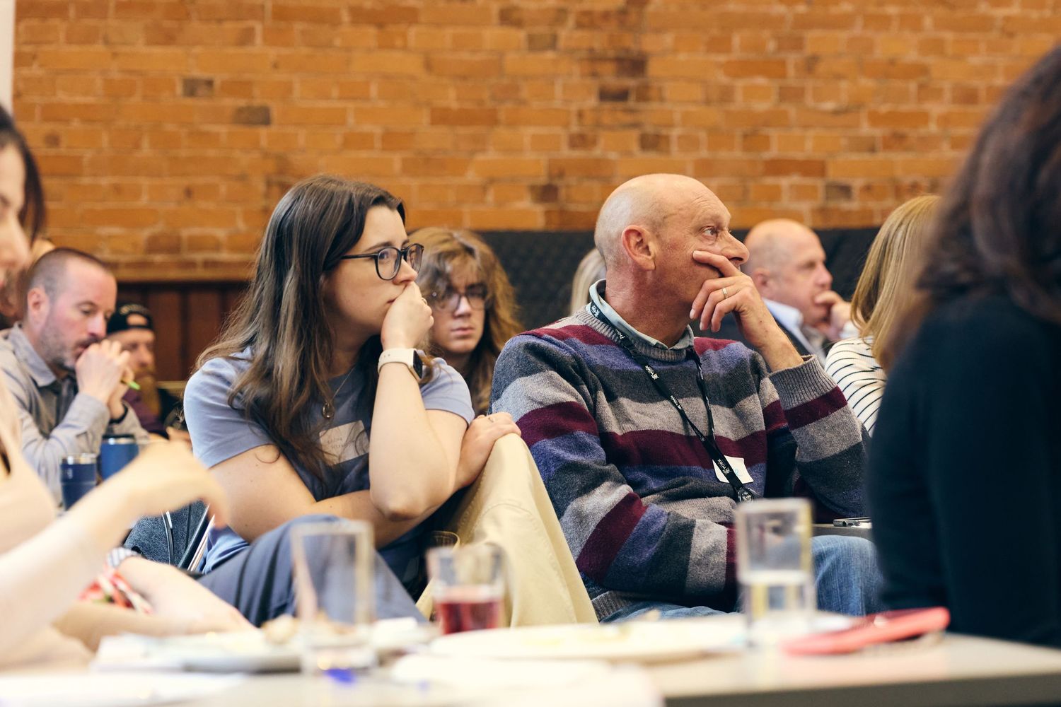 Audience members listening attentively at an indoor conference event, seated at tables in a brick-walled venue.