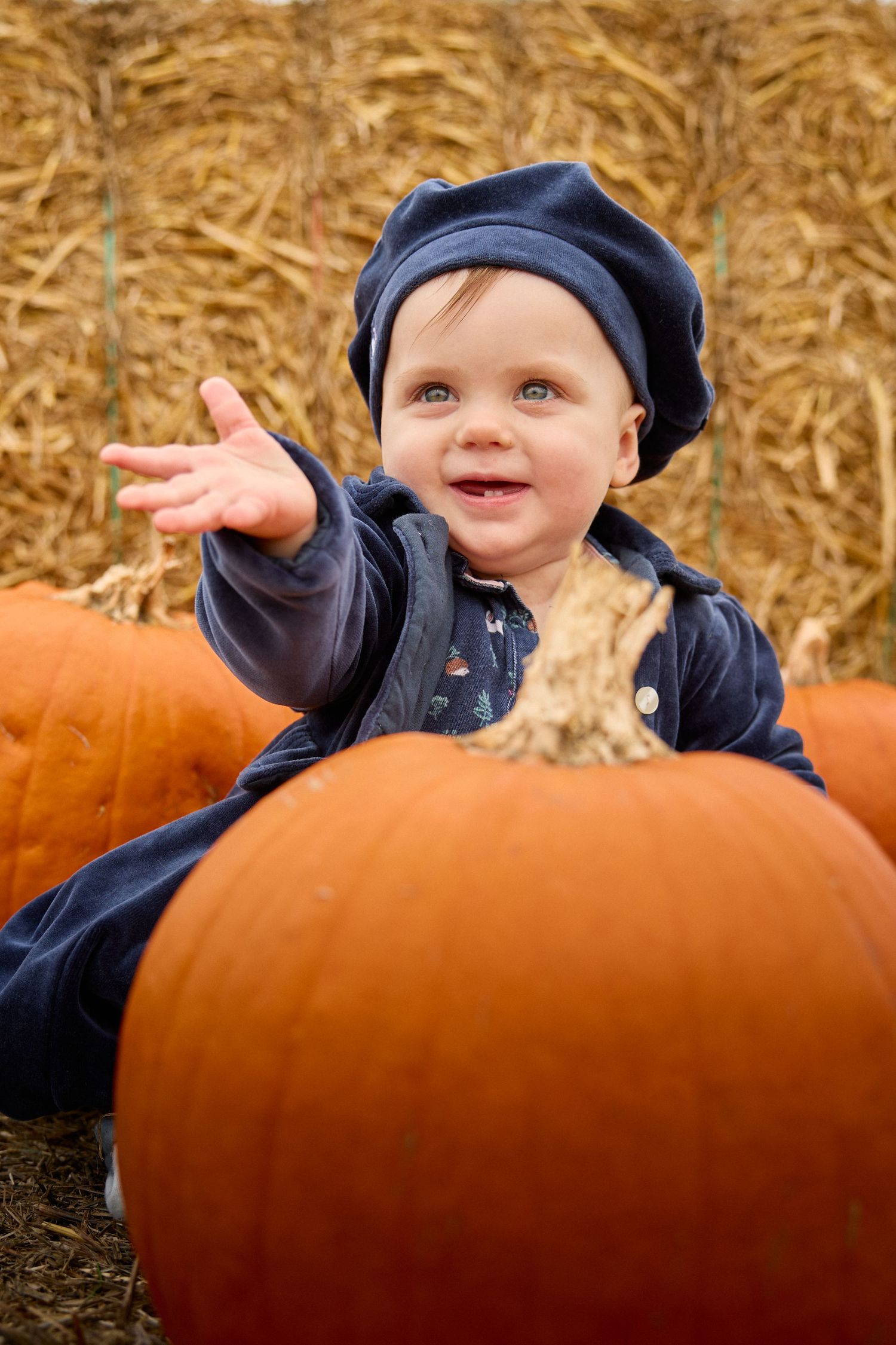 A smiling baby peeks over a large orange pumpkin in an autumn field.