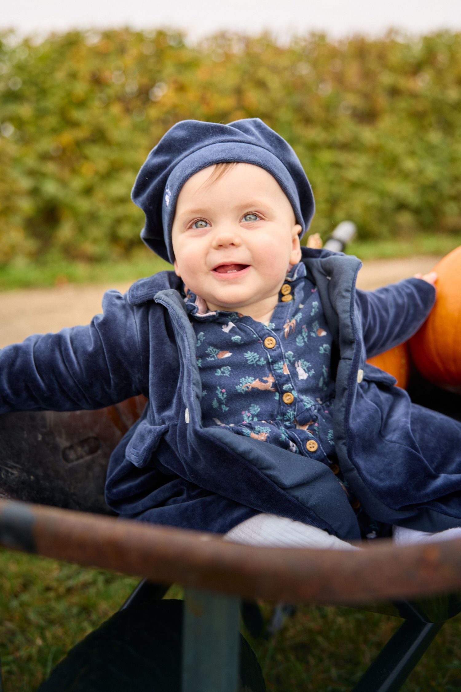 A baby in a navy outfit and hat lies on a dark surface while reaching out playfully.