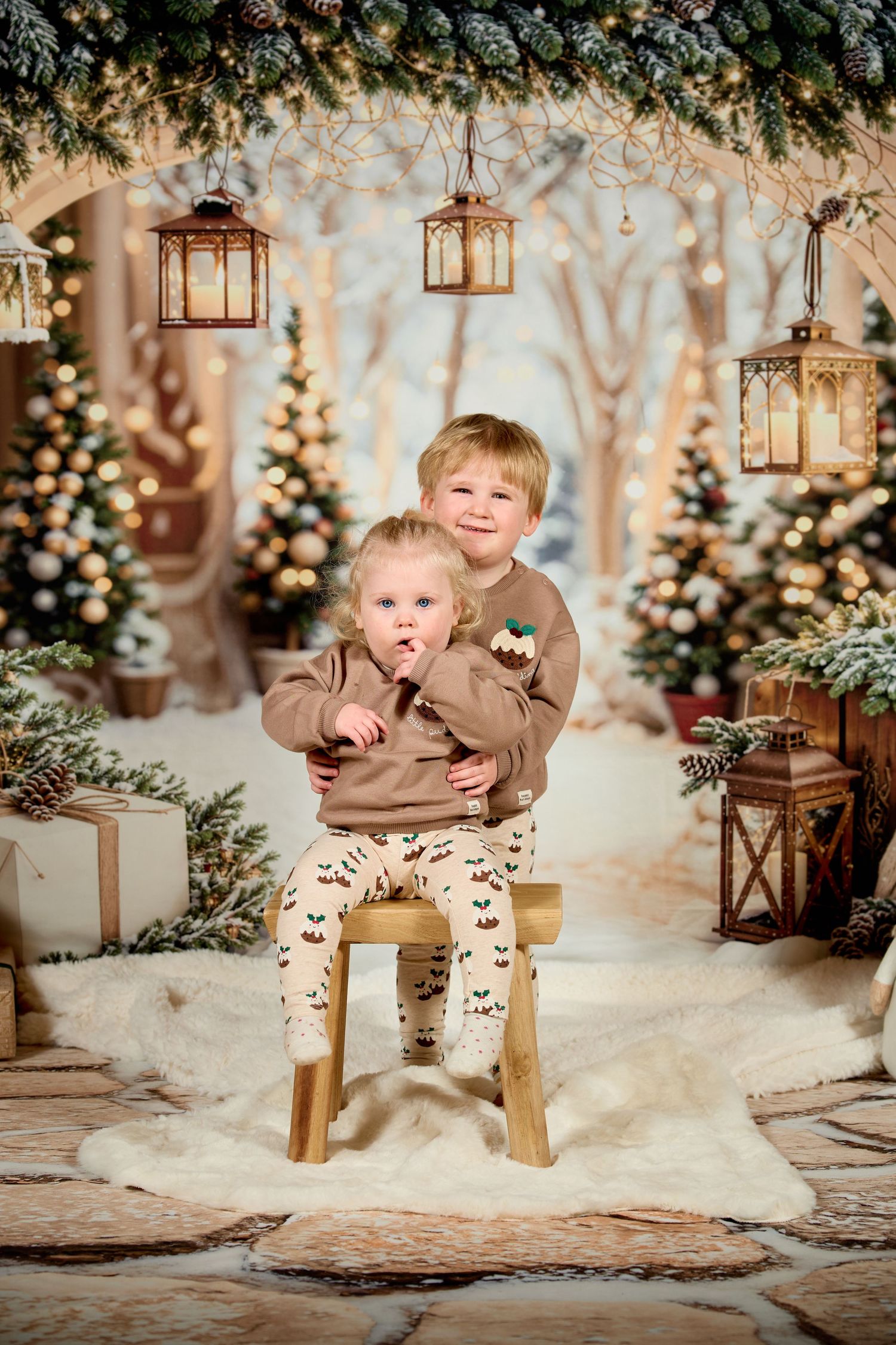 Festive winter photo session with rustic lanterns, Christmas trees, and snow-covered setting in warm golden lighting.