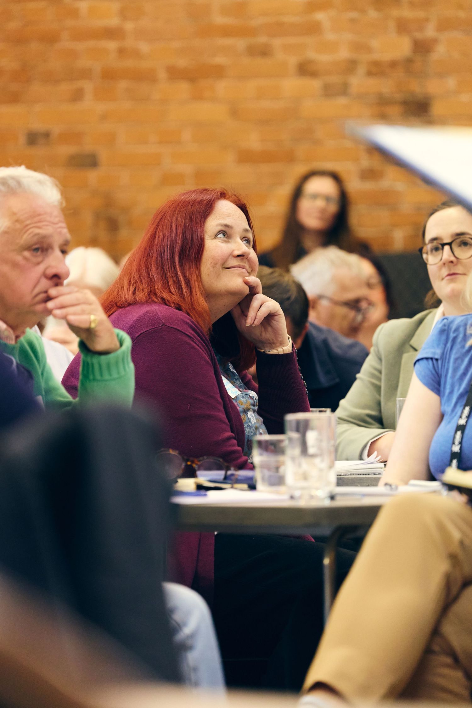 Audience members attentively listening at an indoor seminar, woman with red hair smiling thoughtfully at presentation.