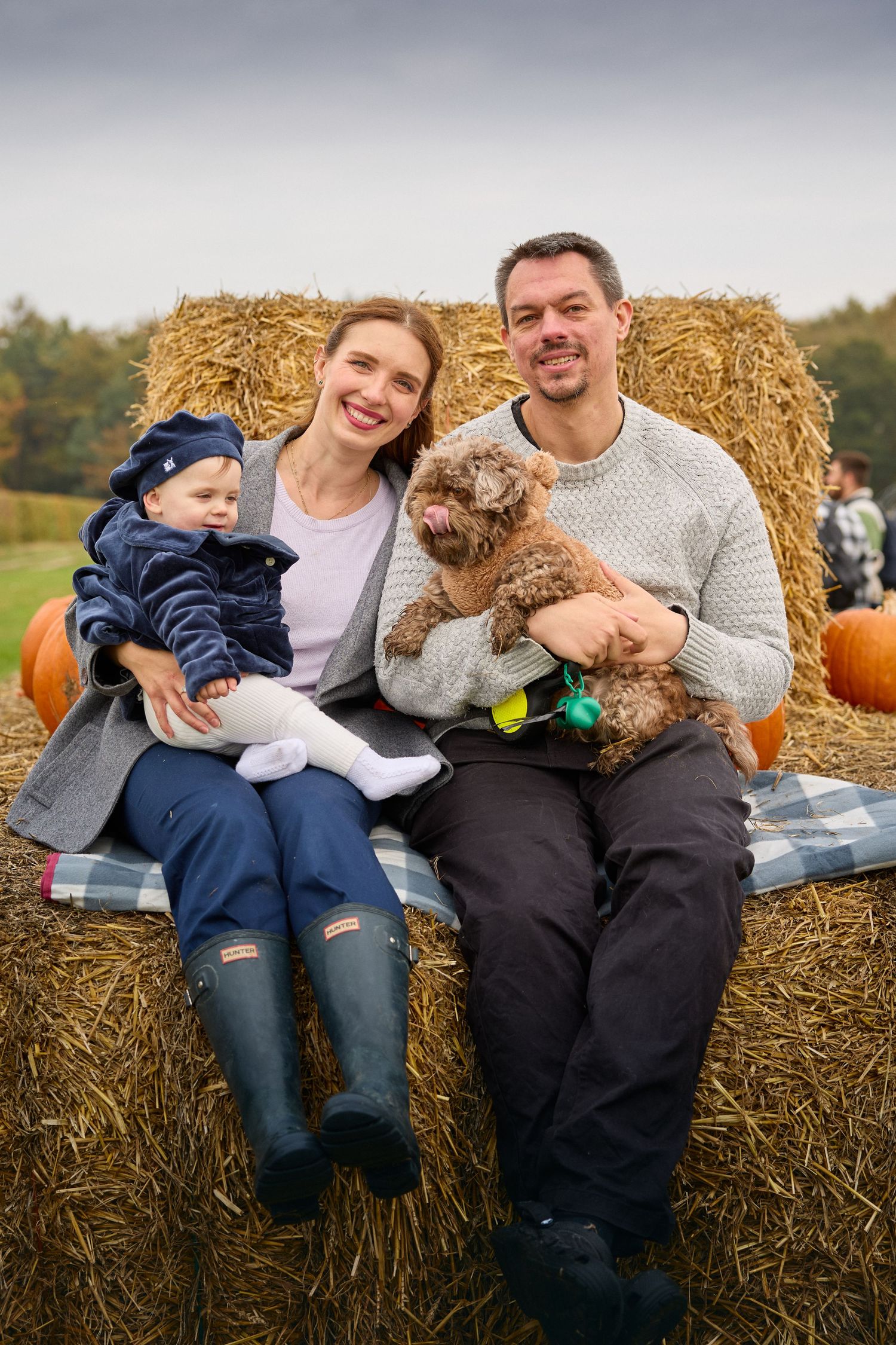 A happy family relaxes on hay bales with their fluffy dog at a pumpkin patch.