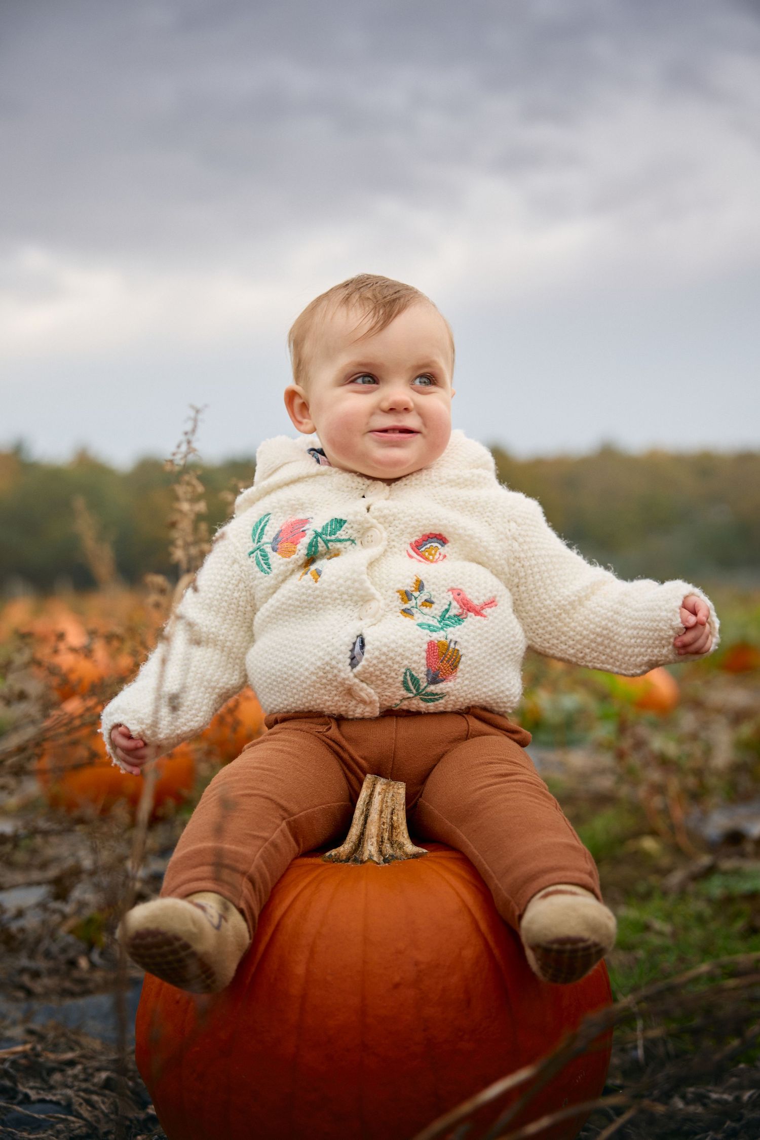 Baby in white embroidered sweater sits on large orange pumpkin under cloudy sky.