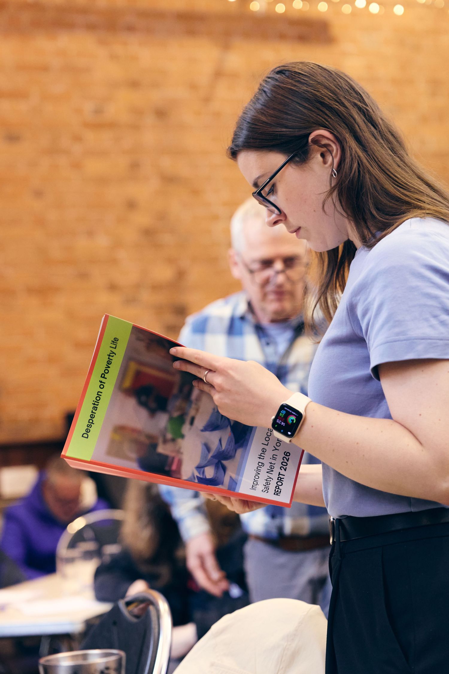 Woman with glasses reads a colorful booklet at an indoor event, wearing an Apple Watch, with brick wall background.