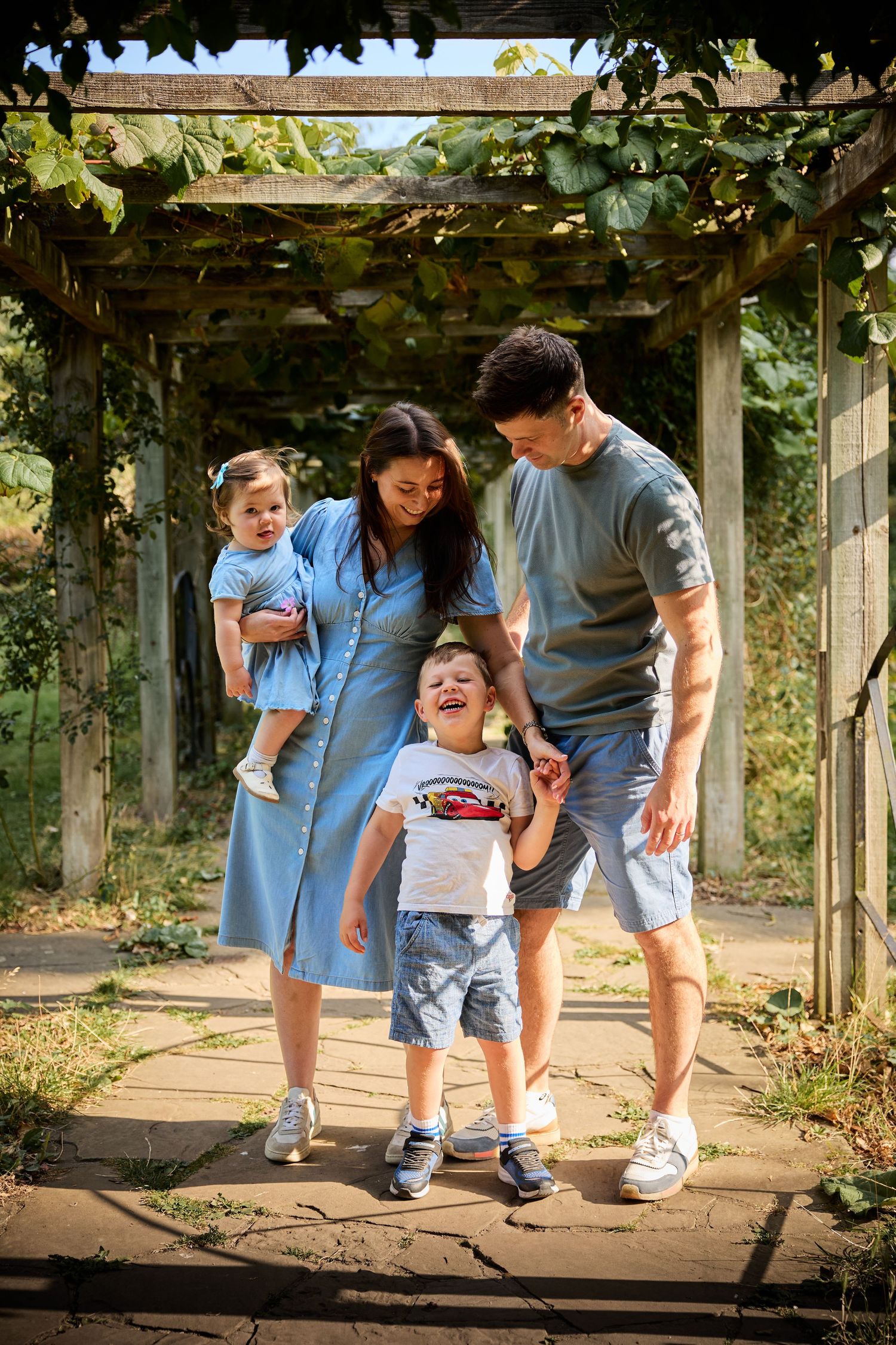 Family walking and playing together on stone path under vine-covered garden arbor.