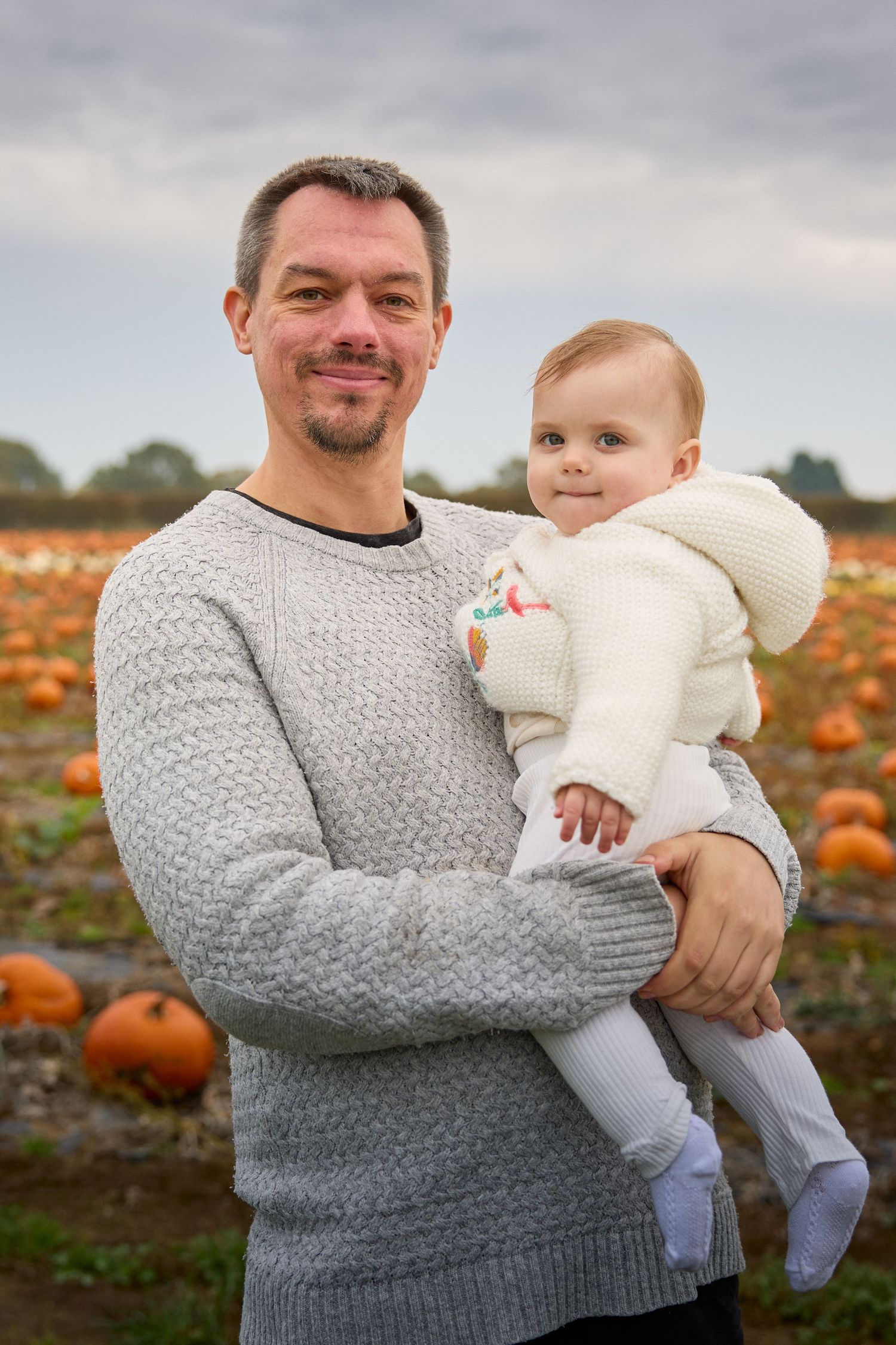 Adult in grey sweater holds baby wearing white outfit in pumpkin patch setting.